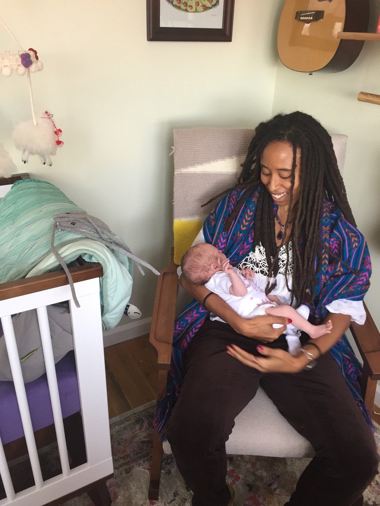 A woman with dreadlocks sitting on a chair, holding a newborn baby in her arms, smiling and looking at the baby. The baby is dressed in white. The background shows a wall with a framed picture, a guitar hanging, and a crib with a mobile hanging above.
