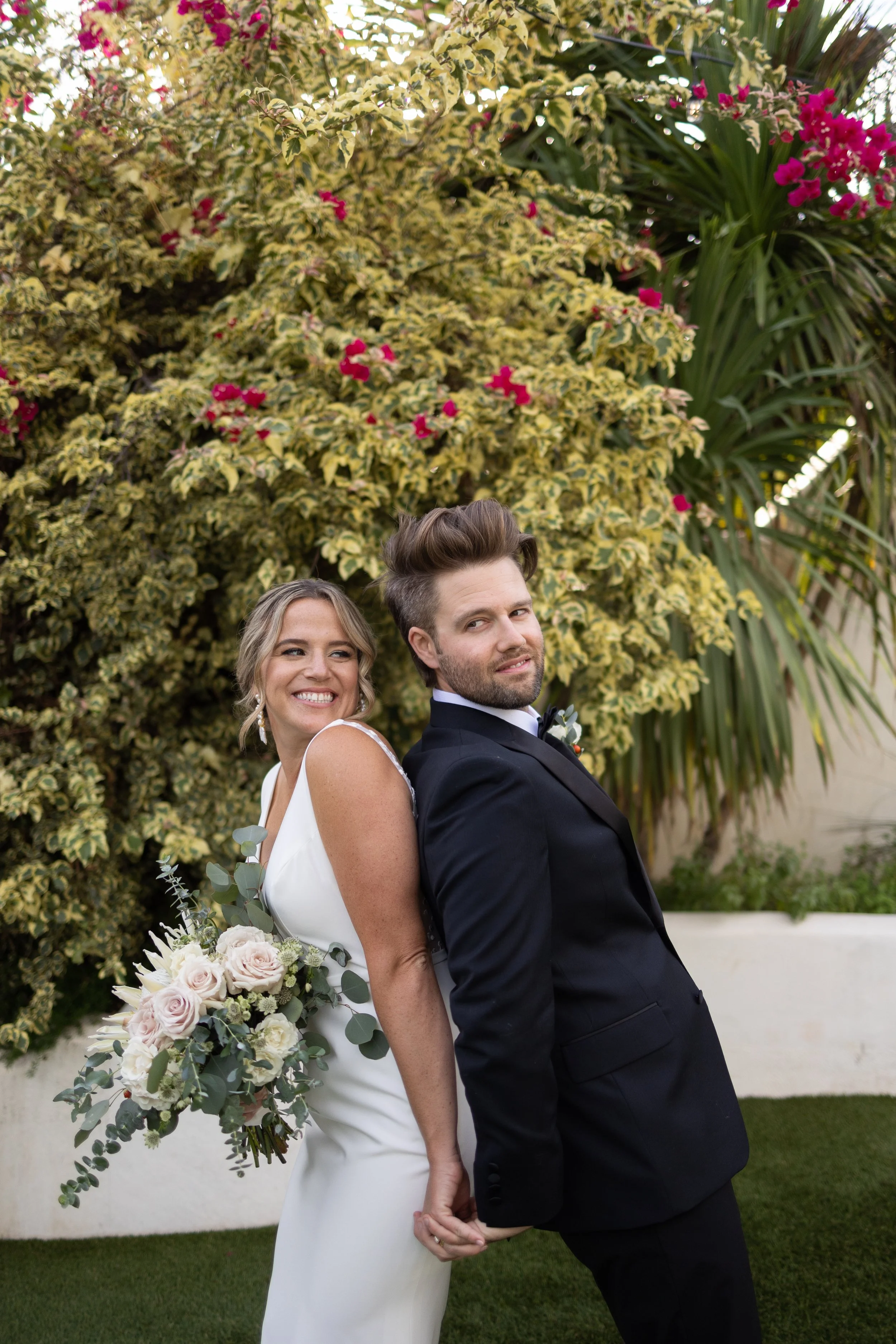 Bride and groom posing playfully back-to-back, bride holding bouquet, in garden setting with flowering shrubs.
