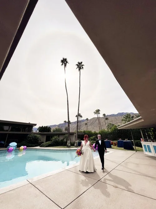A bride and groom walking near a poolside at an outdoor venue with tall palm trees and mountains in the background. The sky is overcast with a halo effect visible around the sun. Colorful inflatables are floating in the pool.