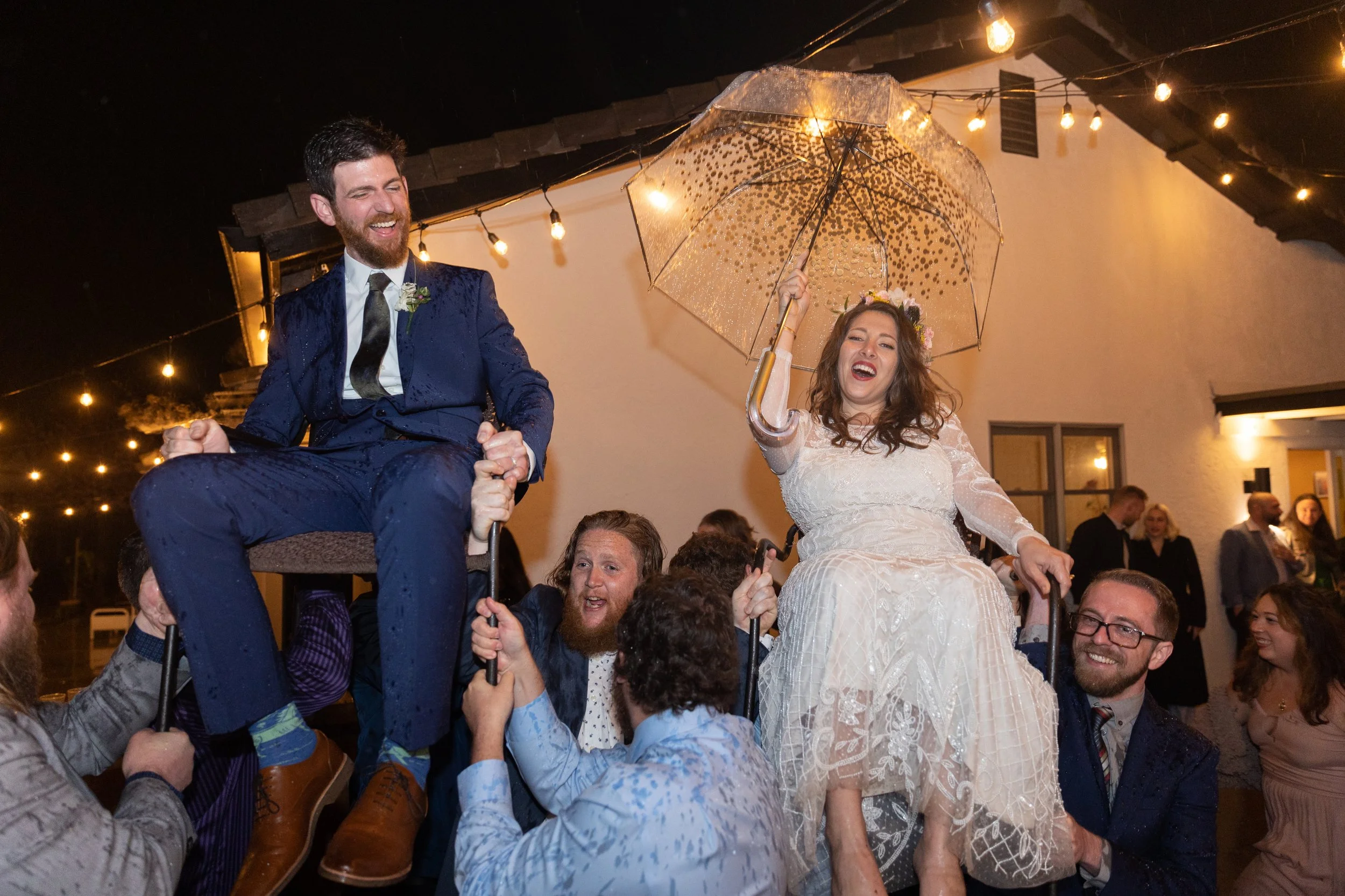 Wedding celebration with a couple being lifted on chairs during the hora dance. The bride holds a transparent umbrella, wearing a white dress, and the groom is in a blue suit. String lights illuminate the outdoor evening setting.