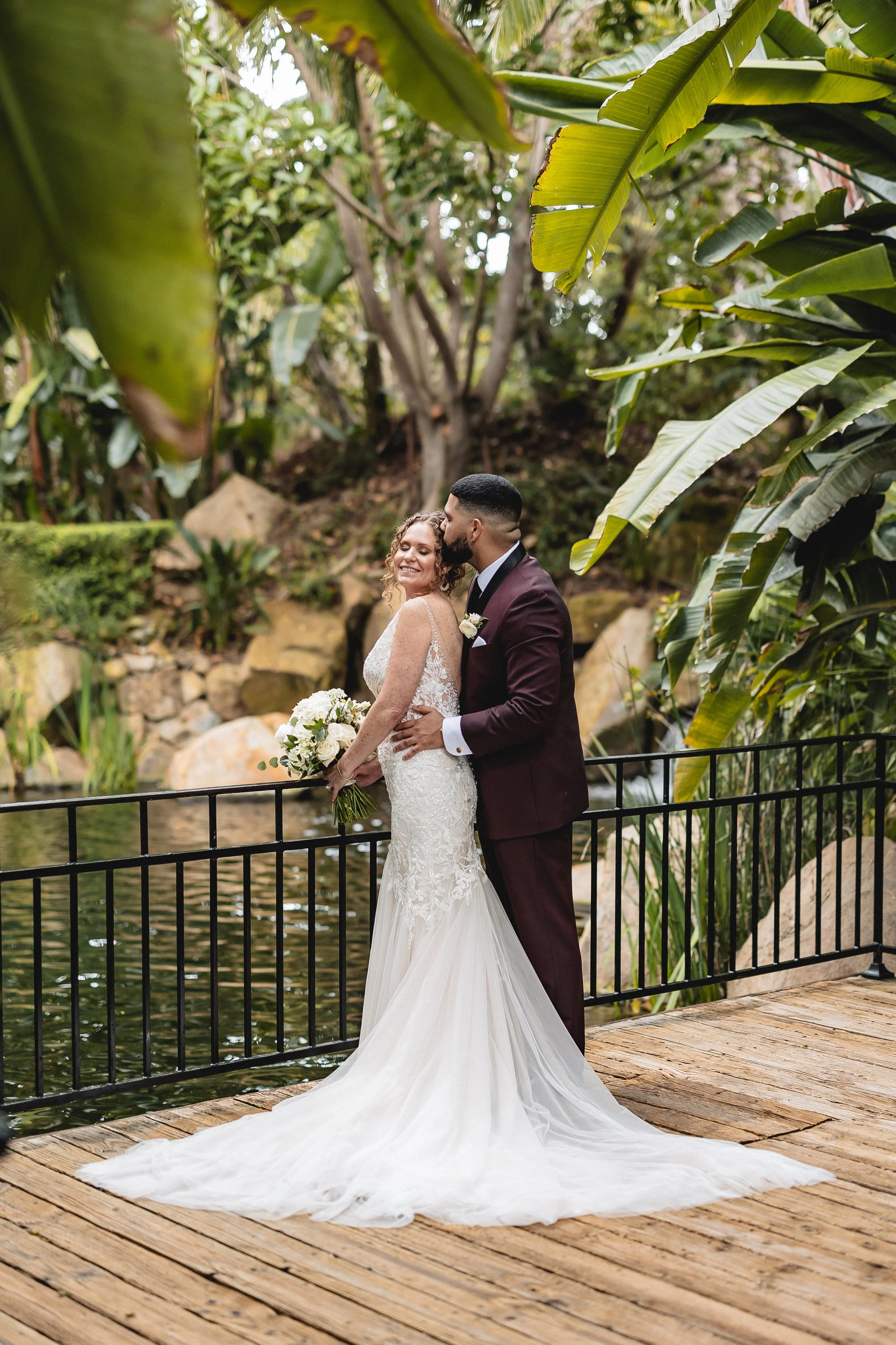Bride in white dress holding bouquet, groom in dark suit embracing, standing by railing near pond, surrounded by lush greenery.