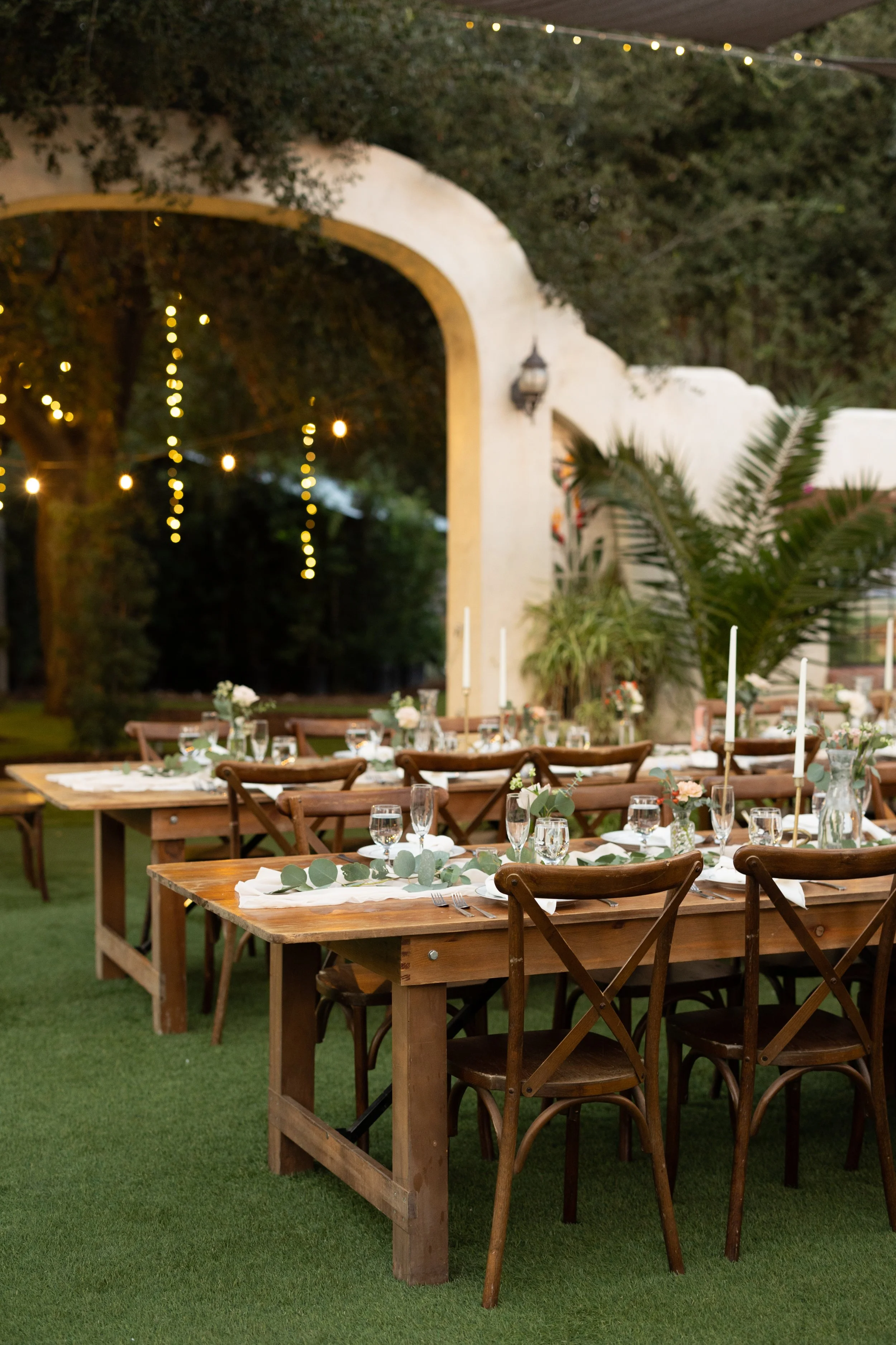 Outdoor wedding reception setup with wooden tables, chairs, and floral decorations, surrounded by greenery and string lights.