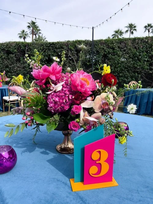 Floral centerpiece on a blue tablecloth with table number 3 at an outdoor event, featuring pink and purple flowers, green foliage, and a purple candle holder, with string lights and trees in the background.