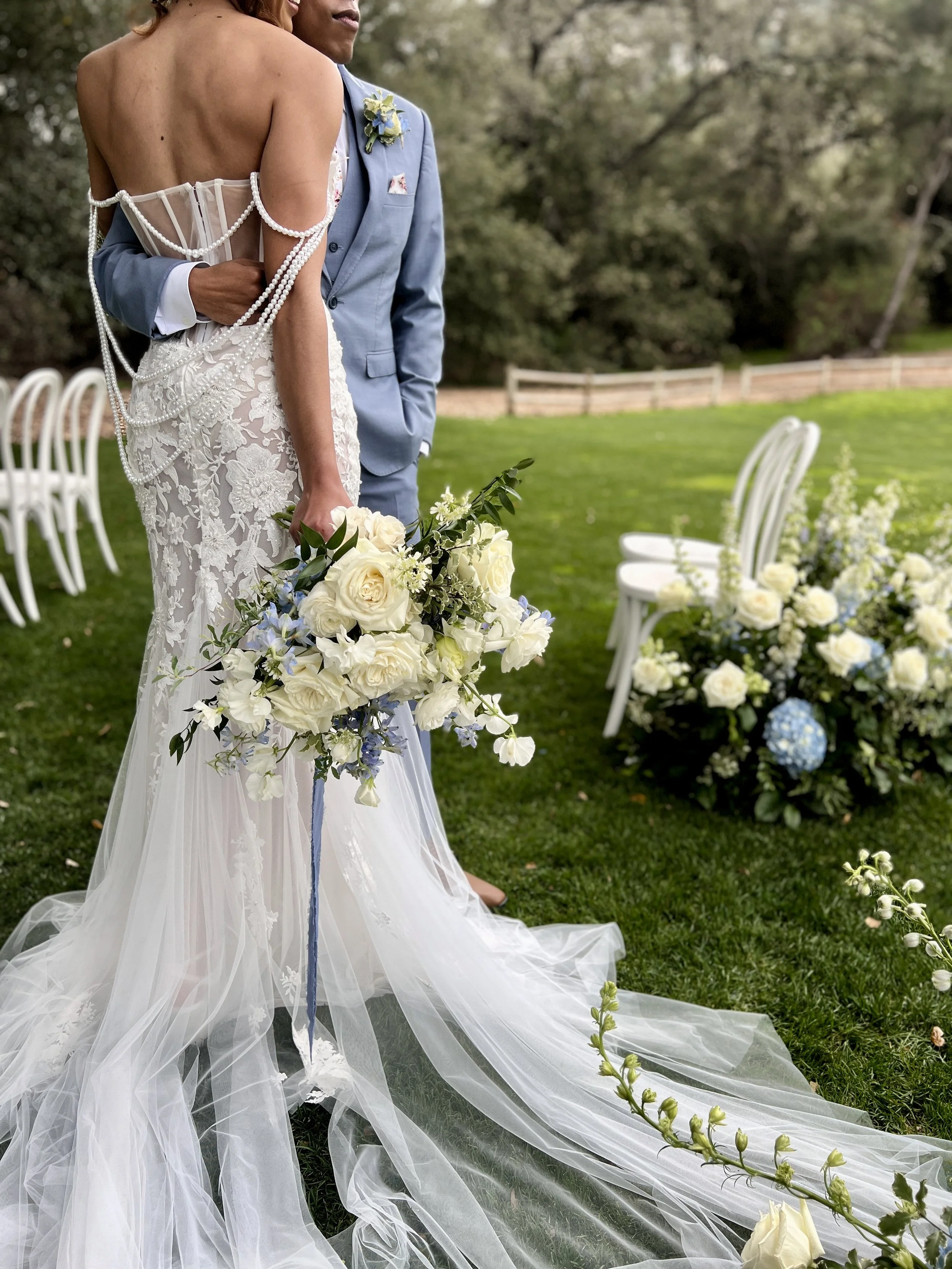 Bride in lace wedding dress holding bouquet while embracing groom in blue suit outdoors.