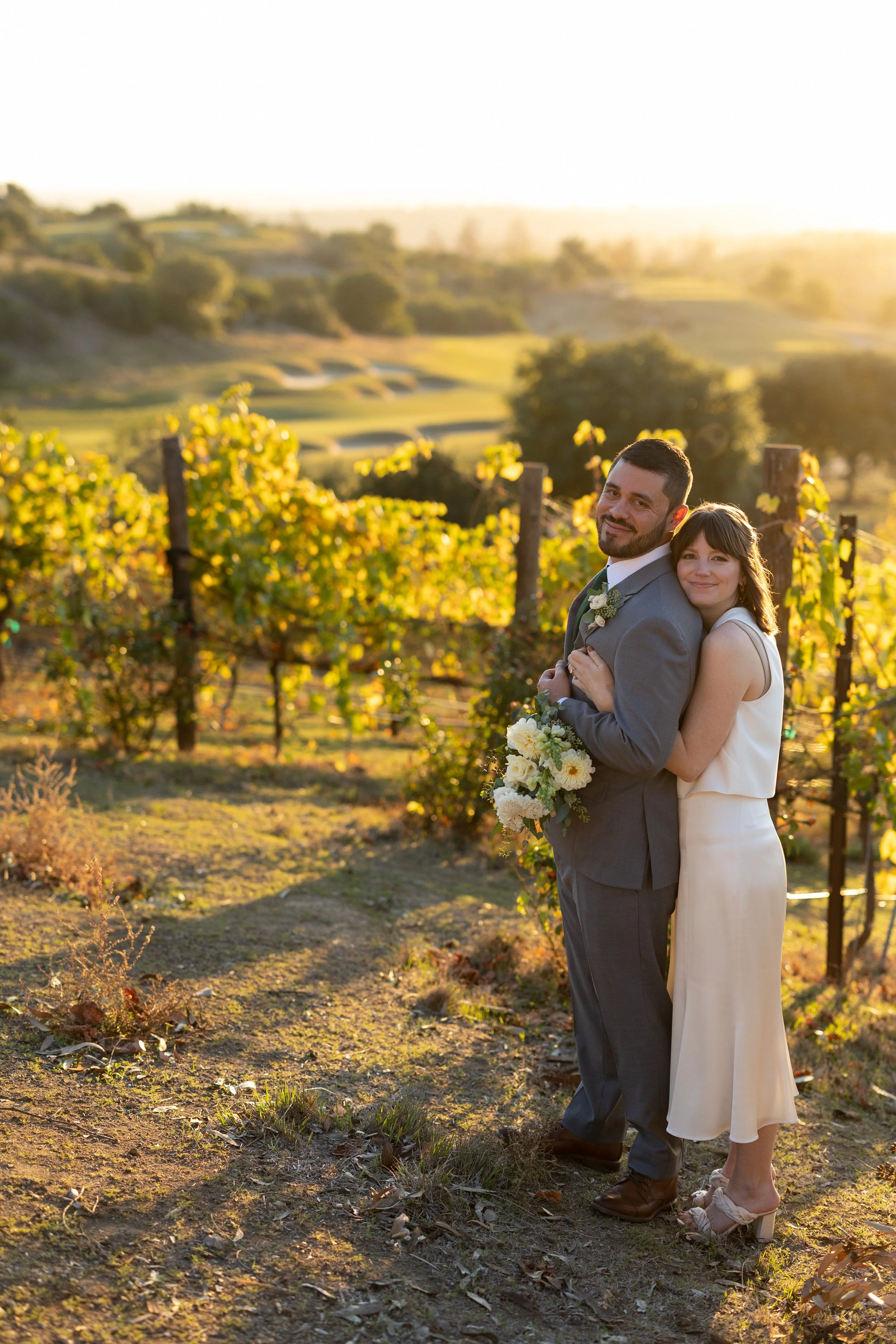 Couple at vineyard during sunset, man in suit, woman in white dress holding bouquet.