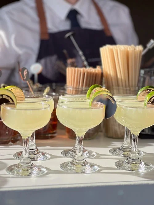 Four margarita cocktails garnished with lime slices at a bar, with bar tools and stirring sticks in the background.