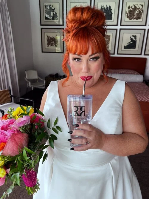 A woman with red hair in an updo wears a white dress, holding a bouquet of colorful flowers in one hand and a clear tumbler with "RR" printed on it in the other. She is sipping from the tumbler through a straw, standing in a room with framed art on t