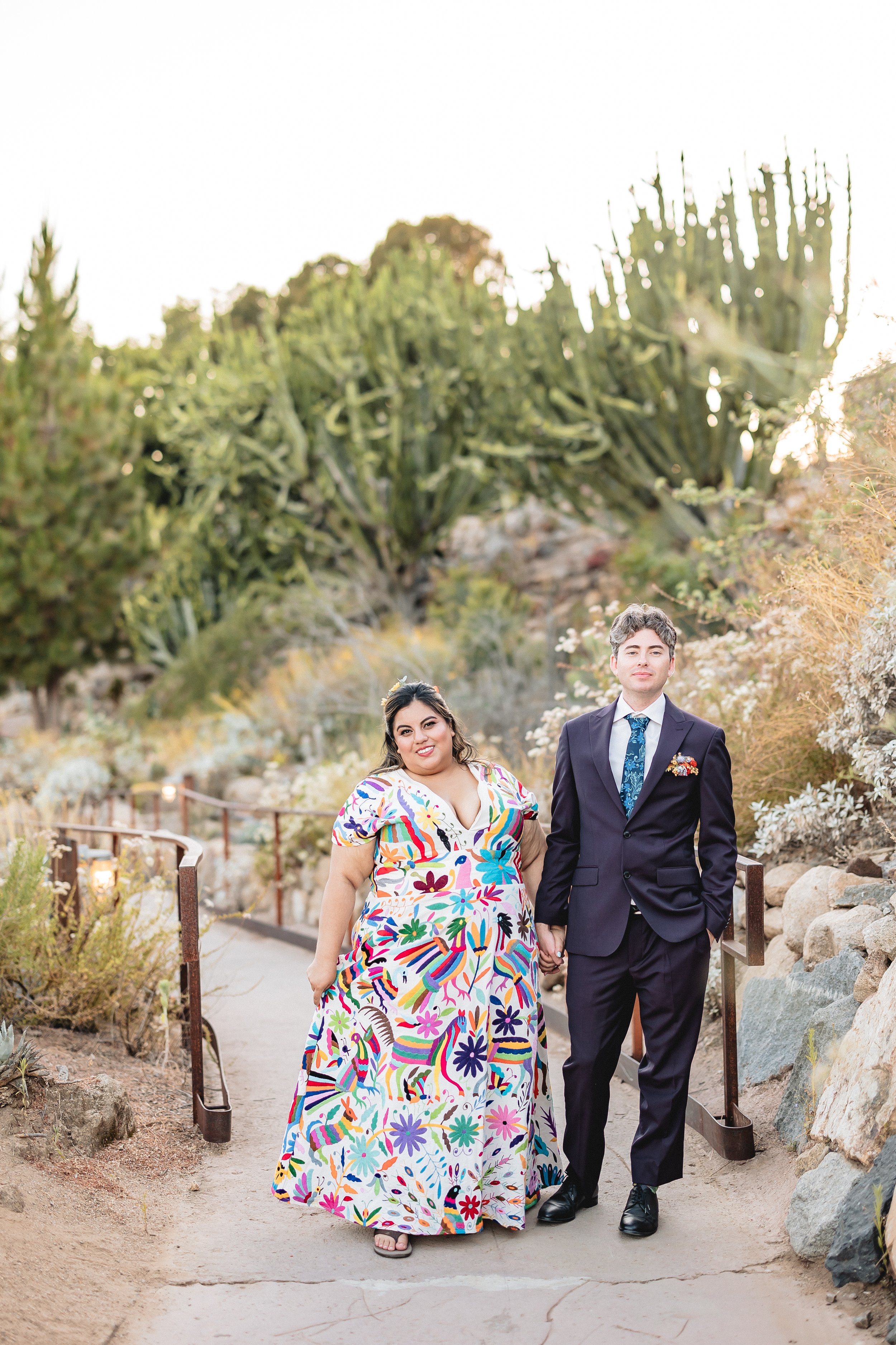 A couple standing on a paved path in a desert garden, with a woman in a colorful floral dress and a man in a suit holding hands. The background features tall cacti and shrubs.