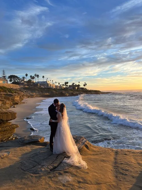 Bride and groom embracing on a rocky beach at sunset with waves and palm trees in the background.