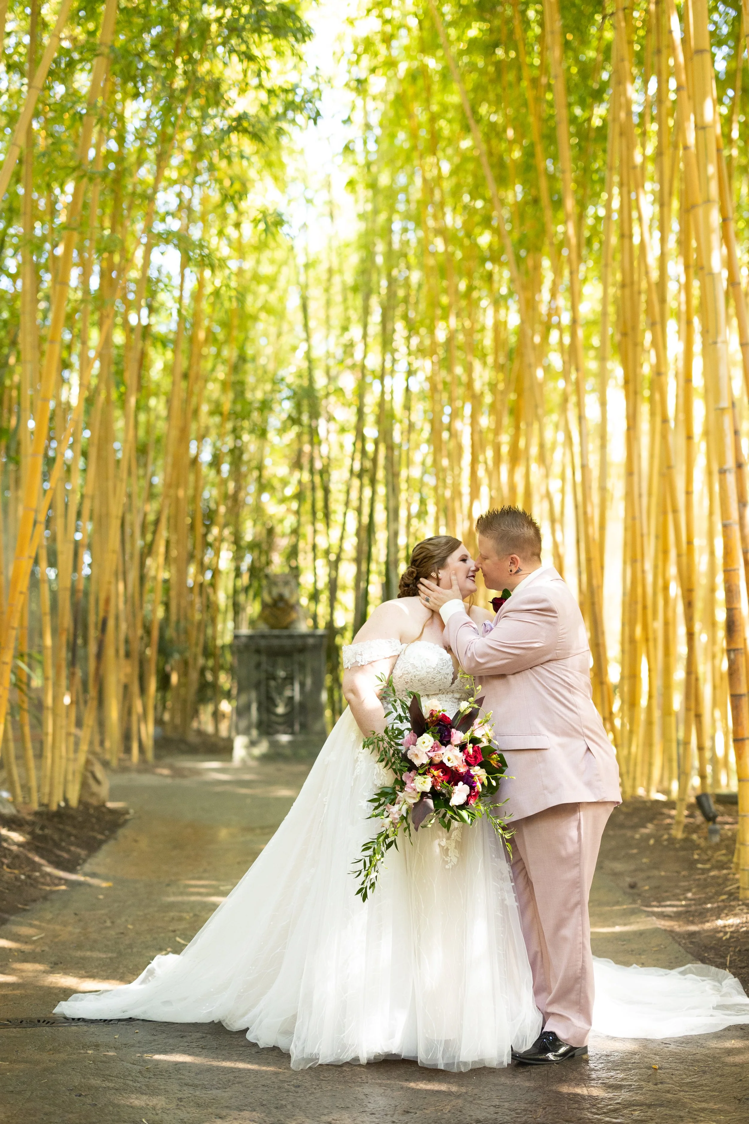 Couple sharing a kiss on their wedding day surrounded by bamboo forest, bride in a white gown with floral bouquet, groom in light pink suit.