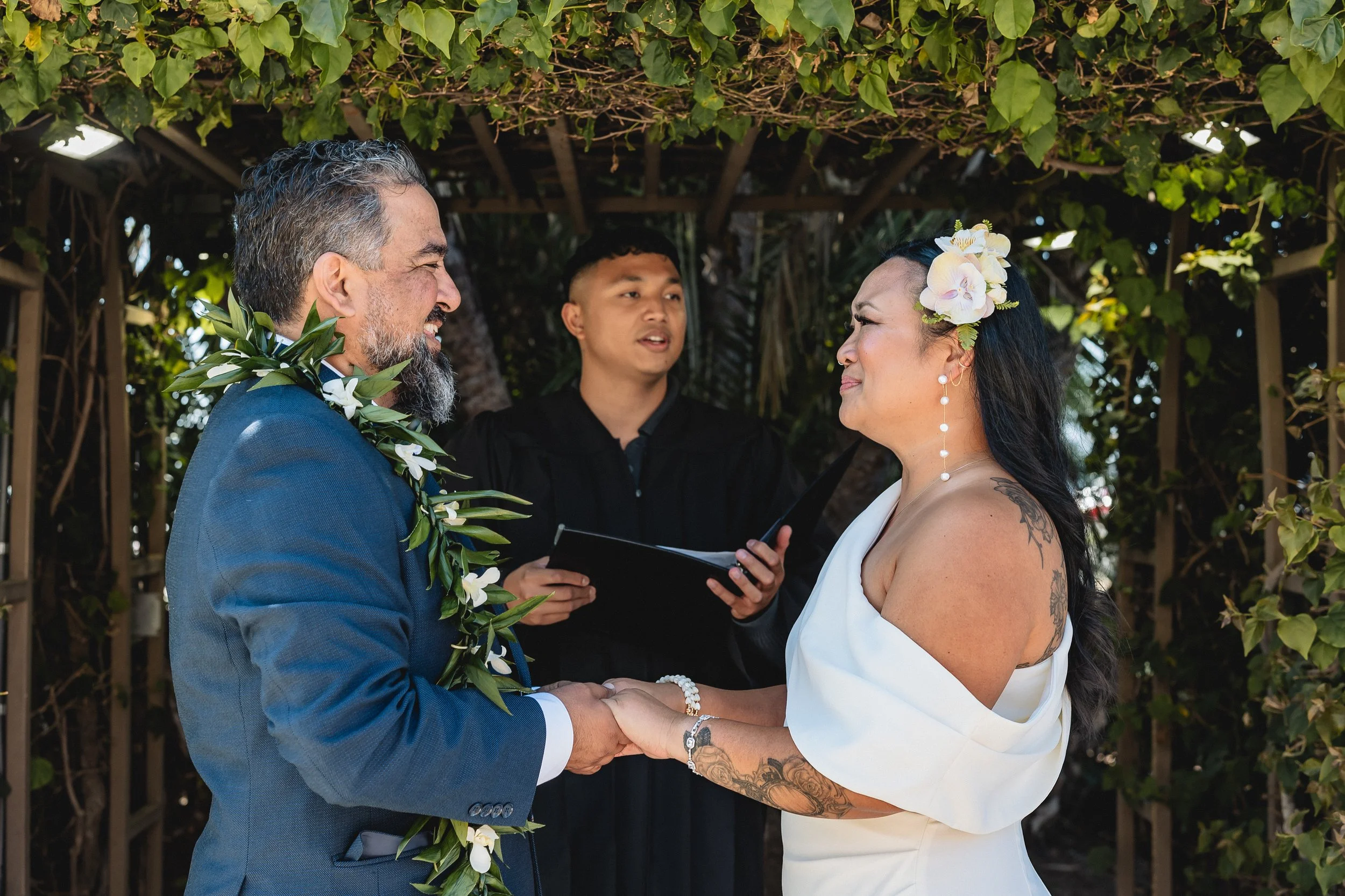 A couple holding hands during a wedding ceremony outdoors, with an officiant standing in the background under a leafy arch. The man is wearing a blue suit and a floral garland, and the woman is in a white dress with a floral hair accessory.