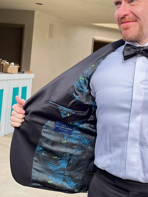 Man wearing a suit with a patterned lining, showing the inside of his jacket. The lining features a blue and black design with a brand label visible. He is dressed in a white shirt and black bow tie.