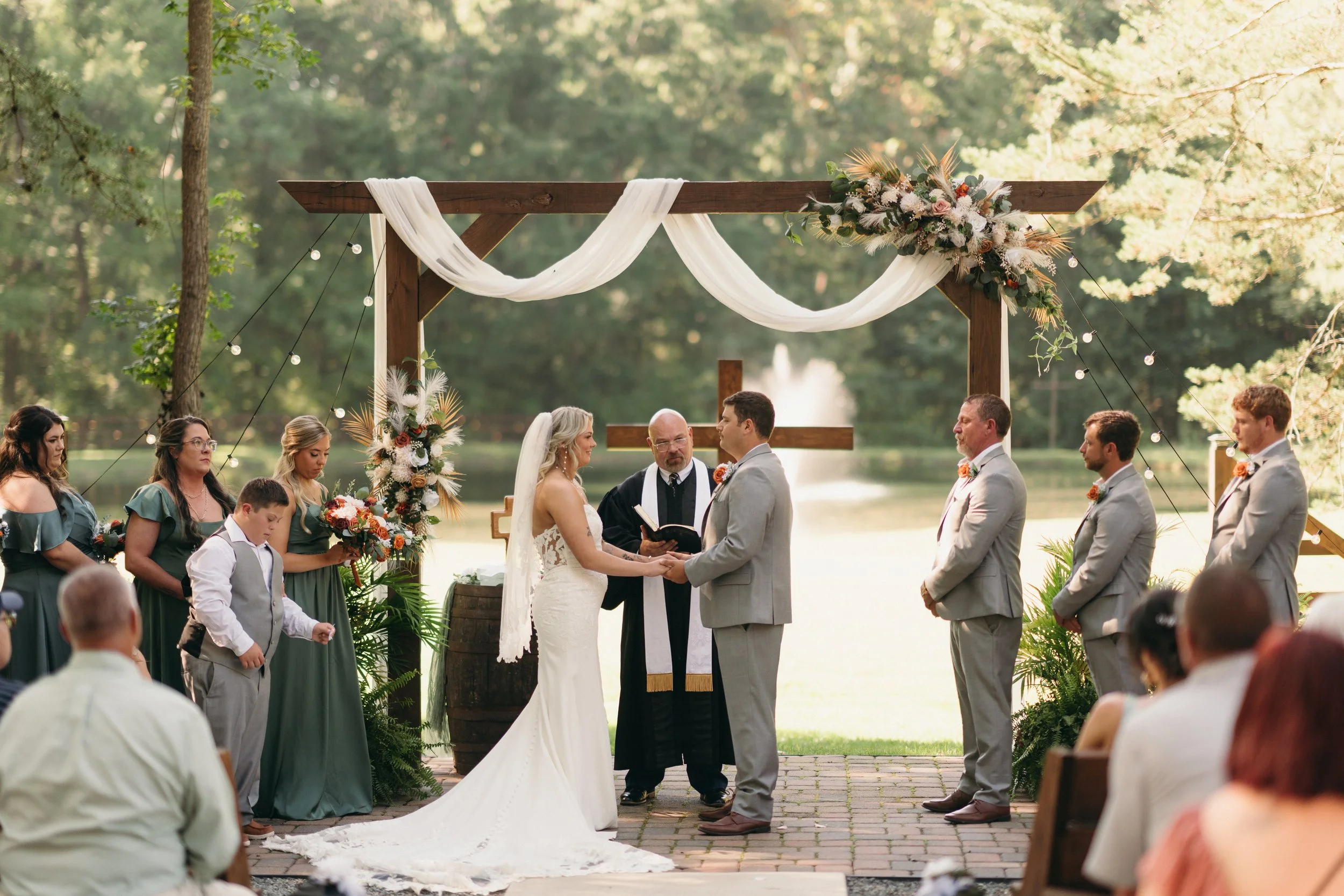 A couple getting married outdoors by a lake, standing under a wooden wedding arch decorated with white drapery and floral arrangements, with friends and family gathered around. Photographer, NC Wedding photographer.