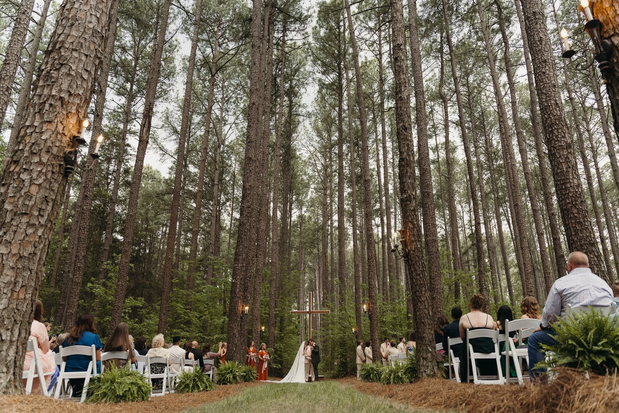 Outdoor wedding ceremony in a forest in NC, with guests seated on white chairs and a cross at the altar, surrounded by tall pine trees.