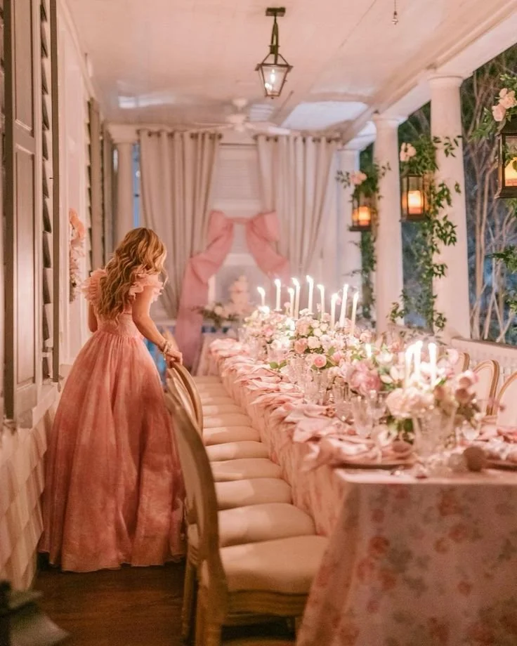 A woman in a pink gown standing at a long, elegantly decorated dining table with pink and white flowers and lit candles inside a beautifully decorated room.