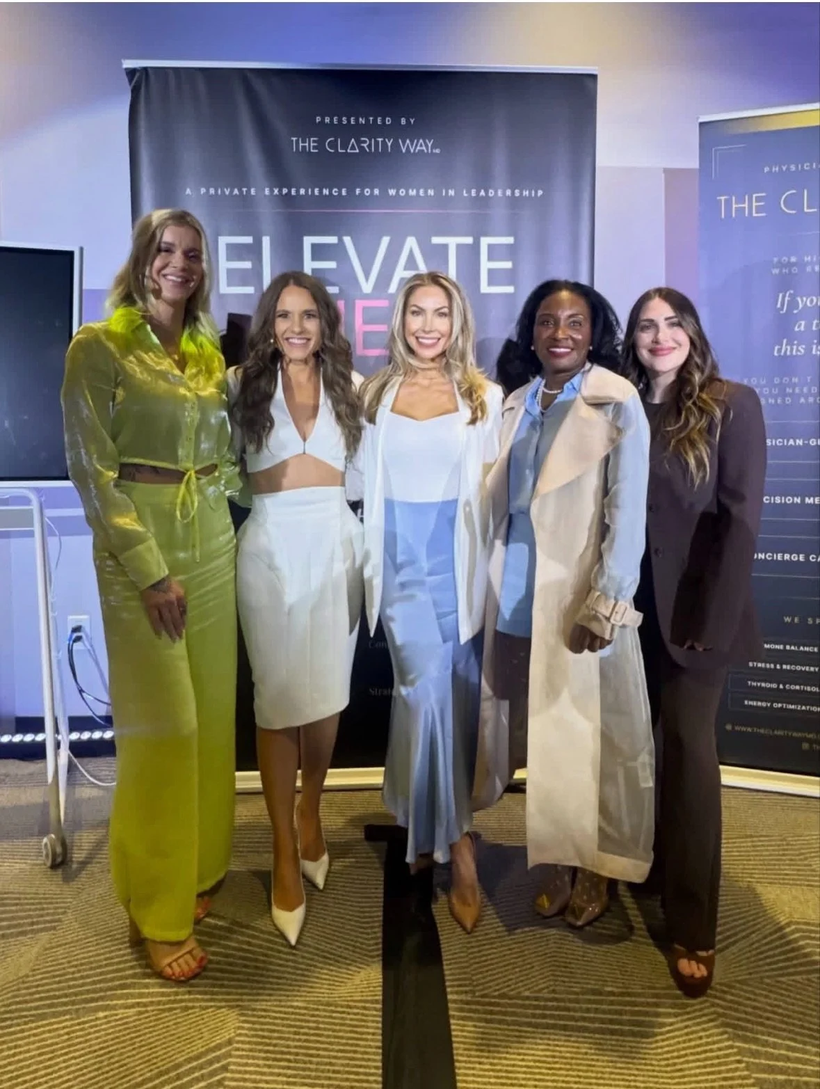 Group of five women standing together at an event, smiling, in front of a banner that reads 'Elevate' and 'Presented by The Clarity Way, A Private Experience for Women in Leadership.'