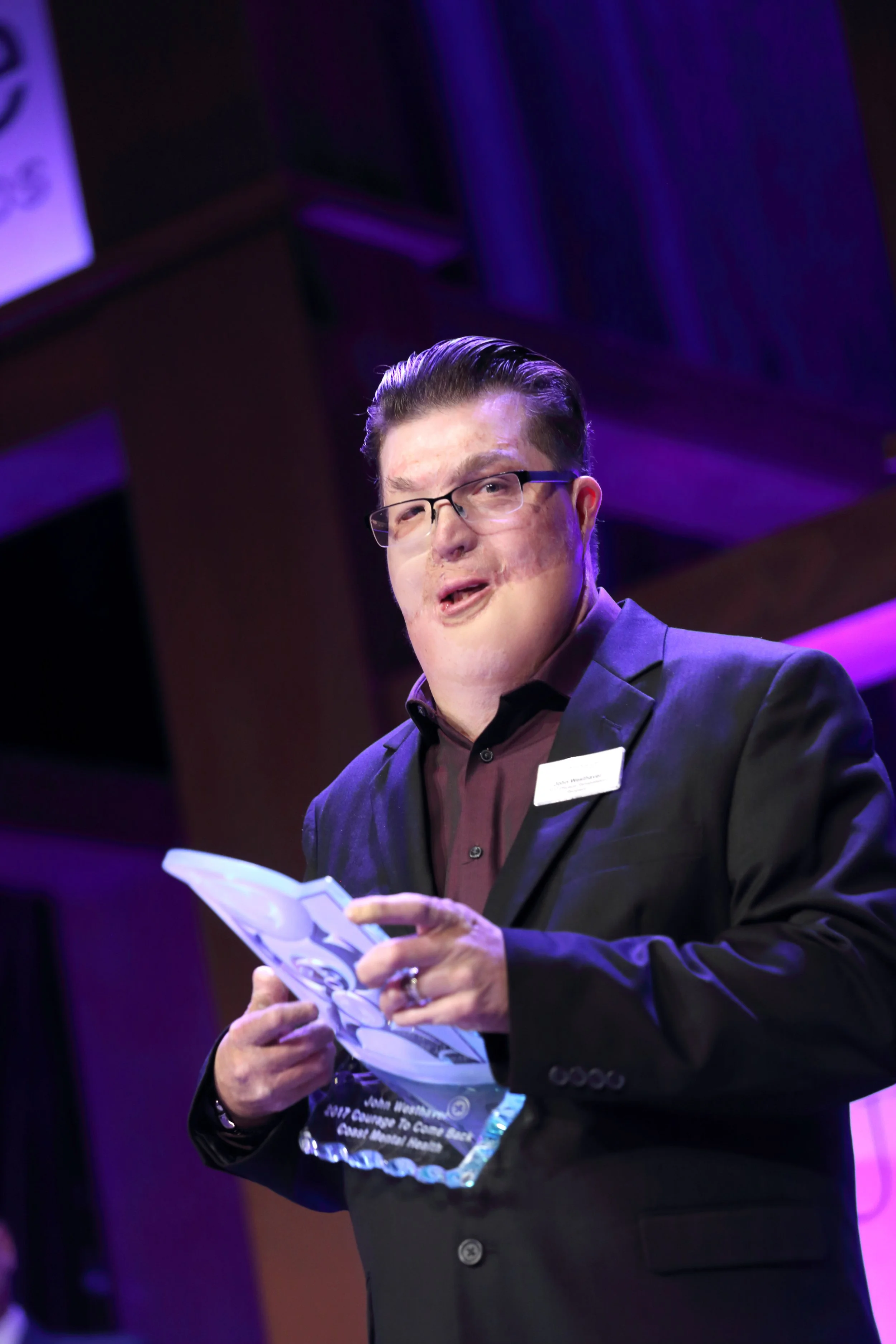 A man in a dark suit and glasses holding an award plaque, speaking at a formal event, with purple lighting in the background.