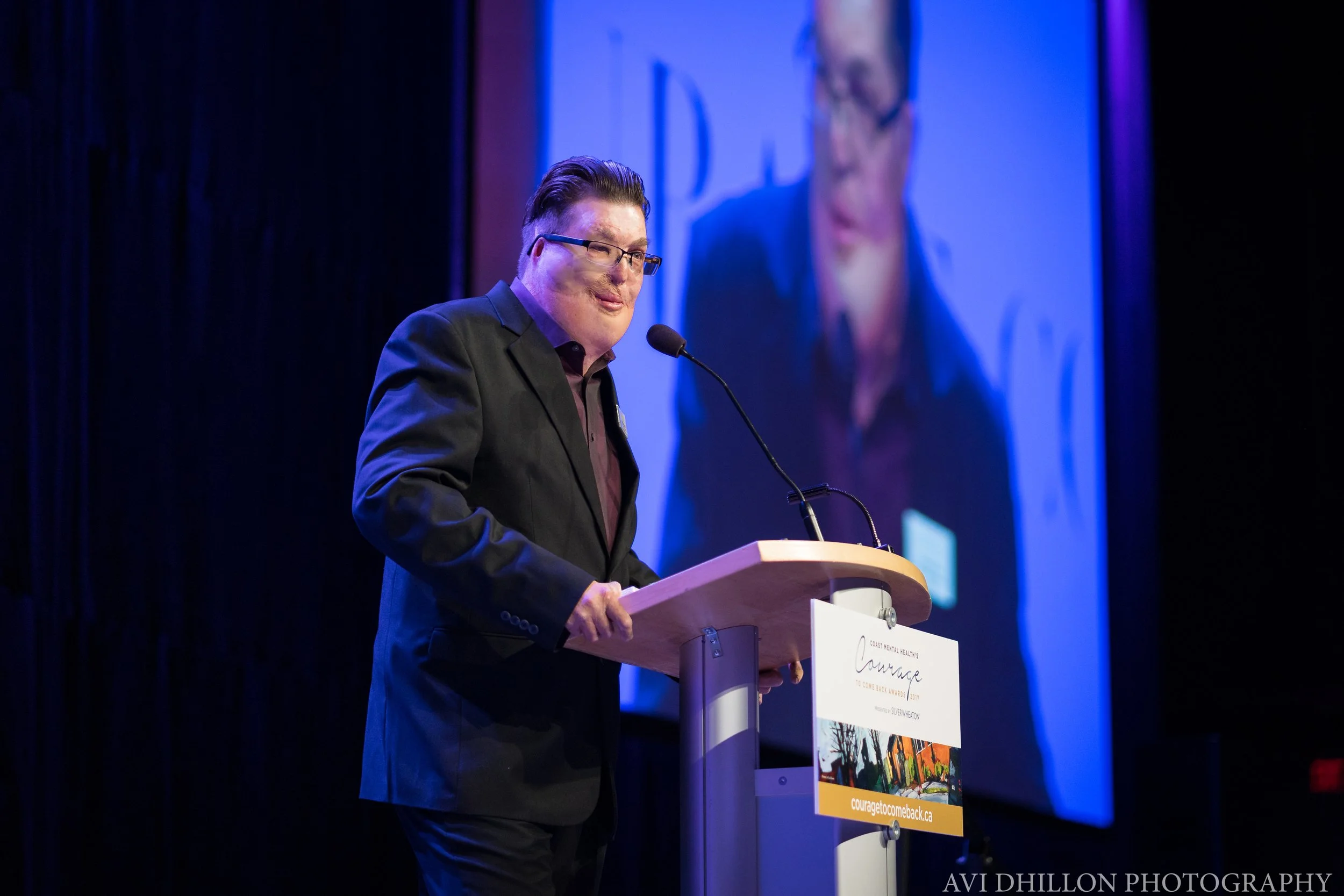 A man in a black suit and glasses standing at a podium speaking into a microphone with a large screen behind him displaying his image.