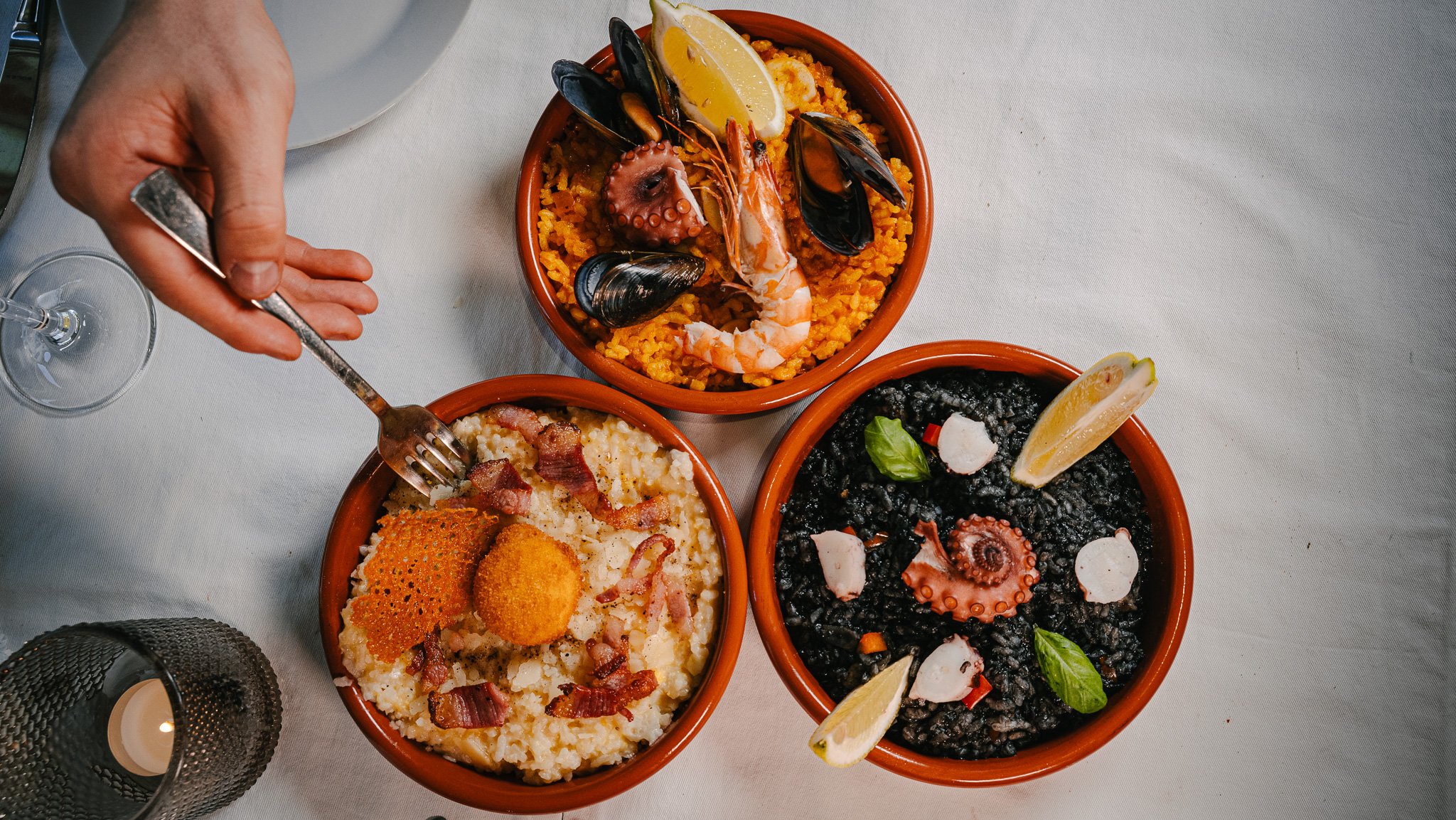 Three bowls of Spanish paella on a white table with a hand holding a fork.
