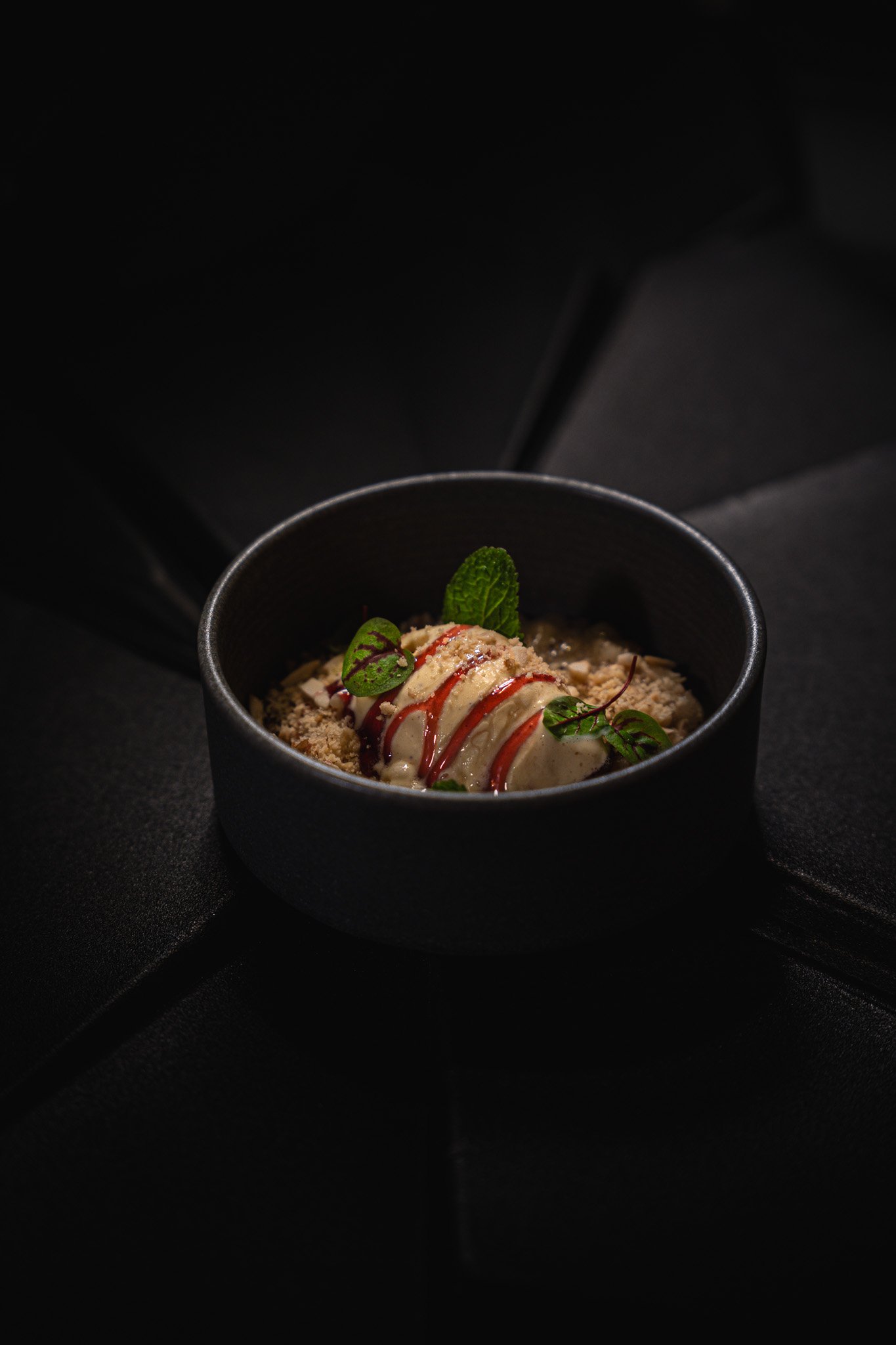 A dark bowl of dessert with garnished mint leaves, a drizzle of red sauce, and sprinkled toppings.