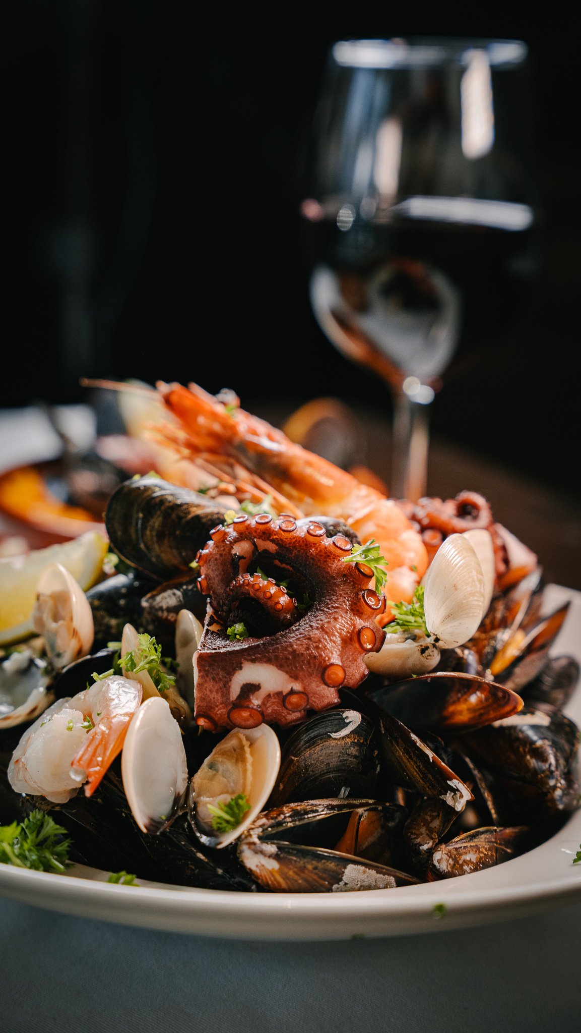 Seafood platter with octopus, shrimp, mussels, and clams, garnished with parsley, with a glass of red wine in the background.