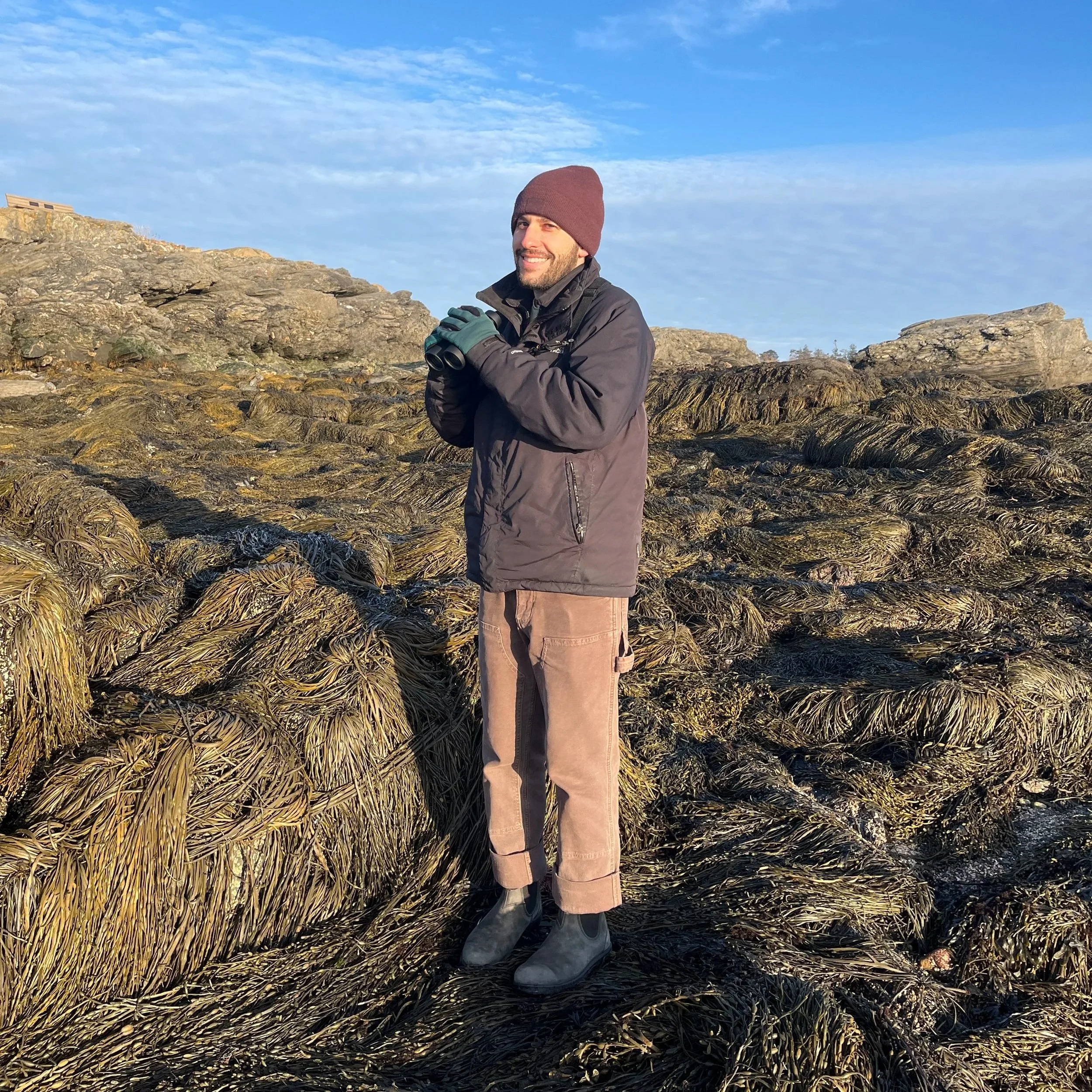 Connor is holding binoculars and smiling while looking towards the sea. The sun is setting in the background.