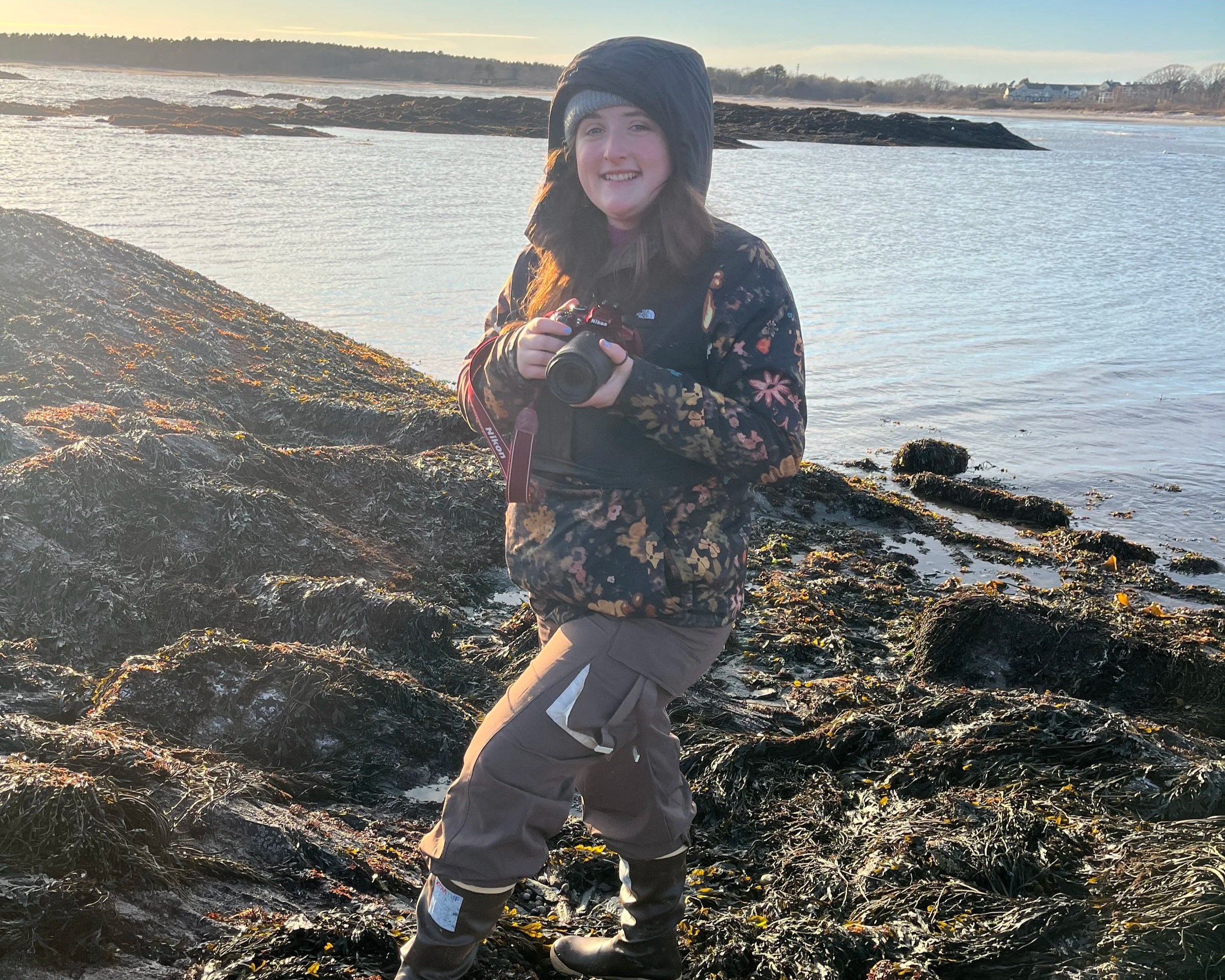 Delia is bundled up in winter clothes and smiling directly at the camera while she holds her own camera.