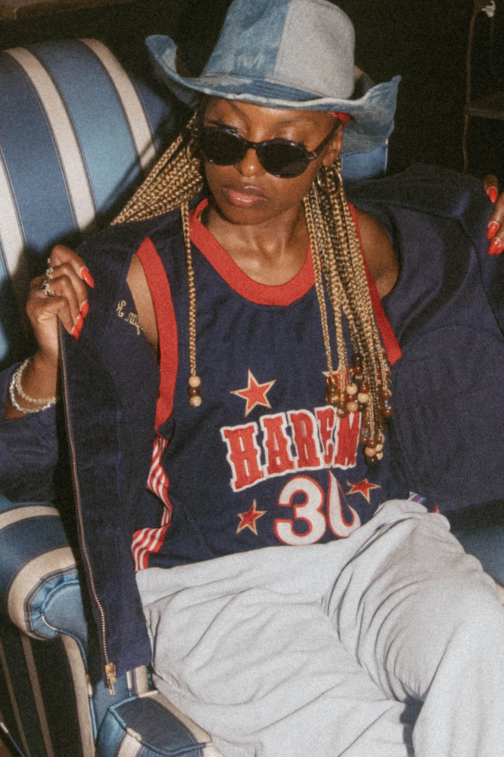 A woman sitting on a blue and white striped sofa, wearing sunglasses, a denim cowboy hat, a navy blue sports jersey with red accents and the number 30, cream-colored pants, and jewelry, with braided hair and red nail polish.