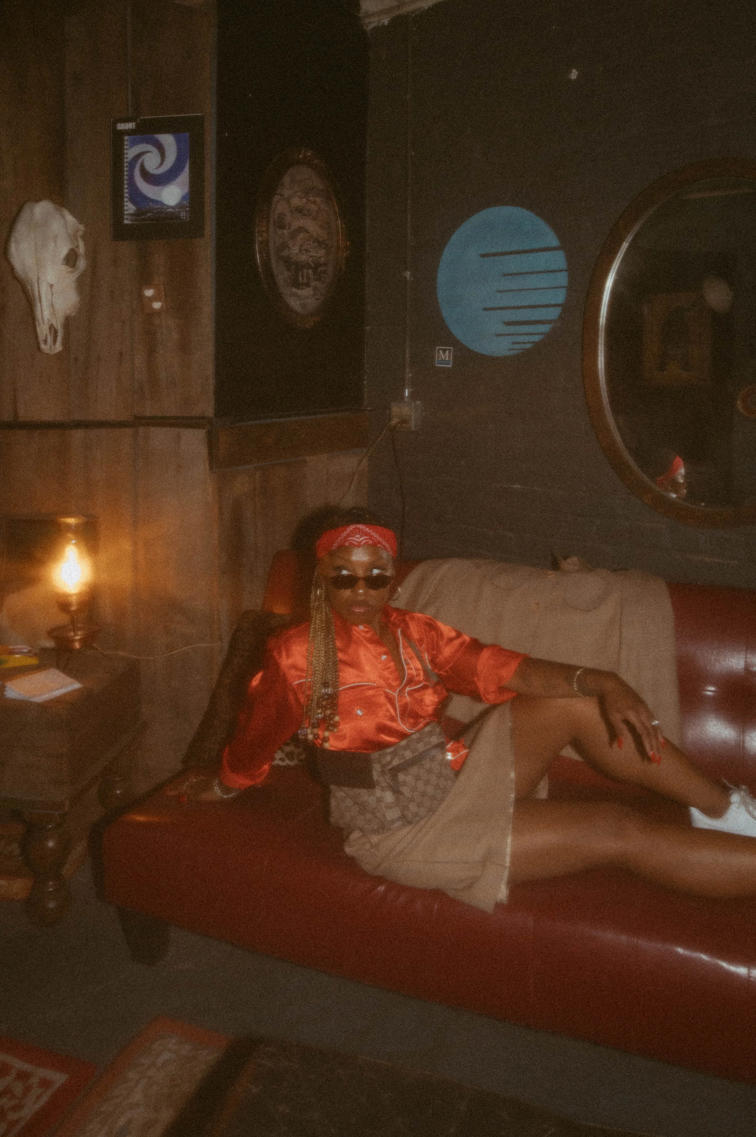 A woman in an orange shirt, beige skirt, and sunglasses sitting on a red leather sofa in a dimly lit room with wood-paneled walls, wall art, and a skull decoration.