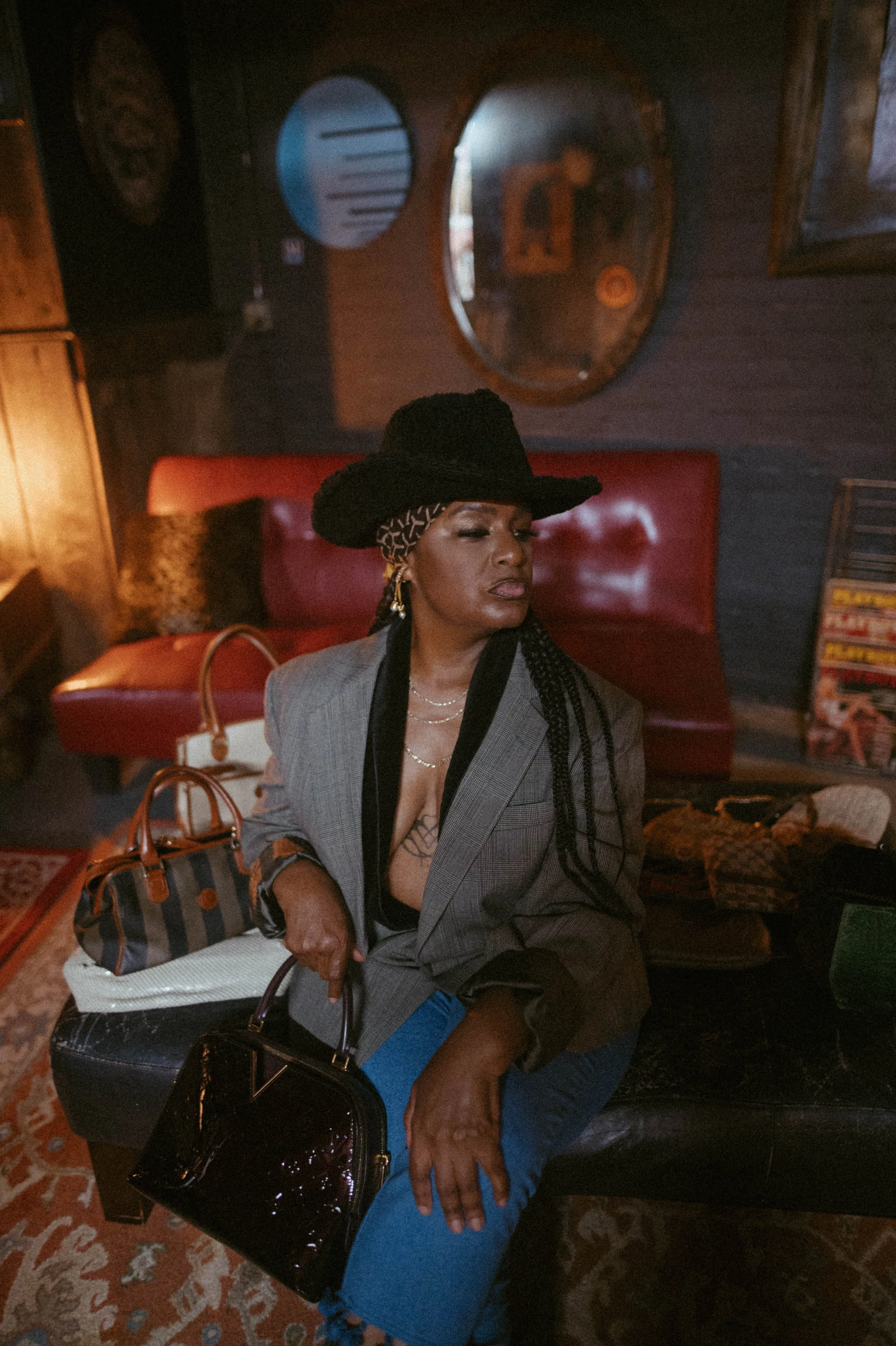 A woman sitting on a black leather bench in a dimly lit room, wearing a gray blazer, blue jeans, and a large black hat, with multiple bags beside her. Behind her is a red upholstered headboard and a dark wooden wall with mirrors and framed artwork.