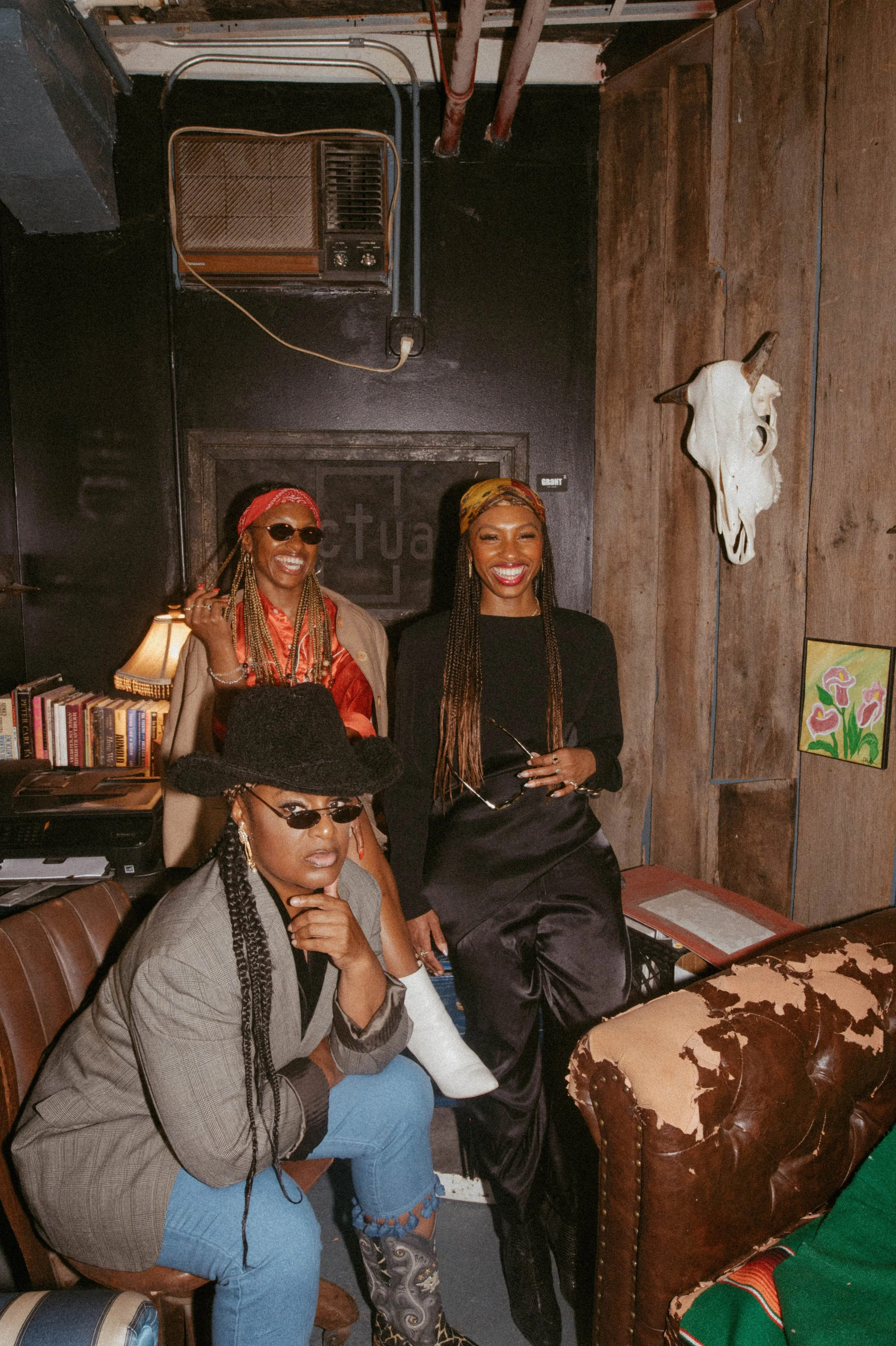 Three Black women posing in a vintage-style study, wearing elevated streetwear including a black cowboy hat, silk wraps, and tailored blazers.