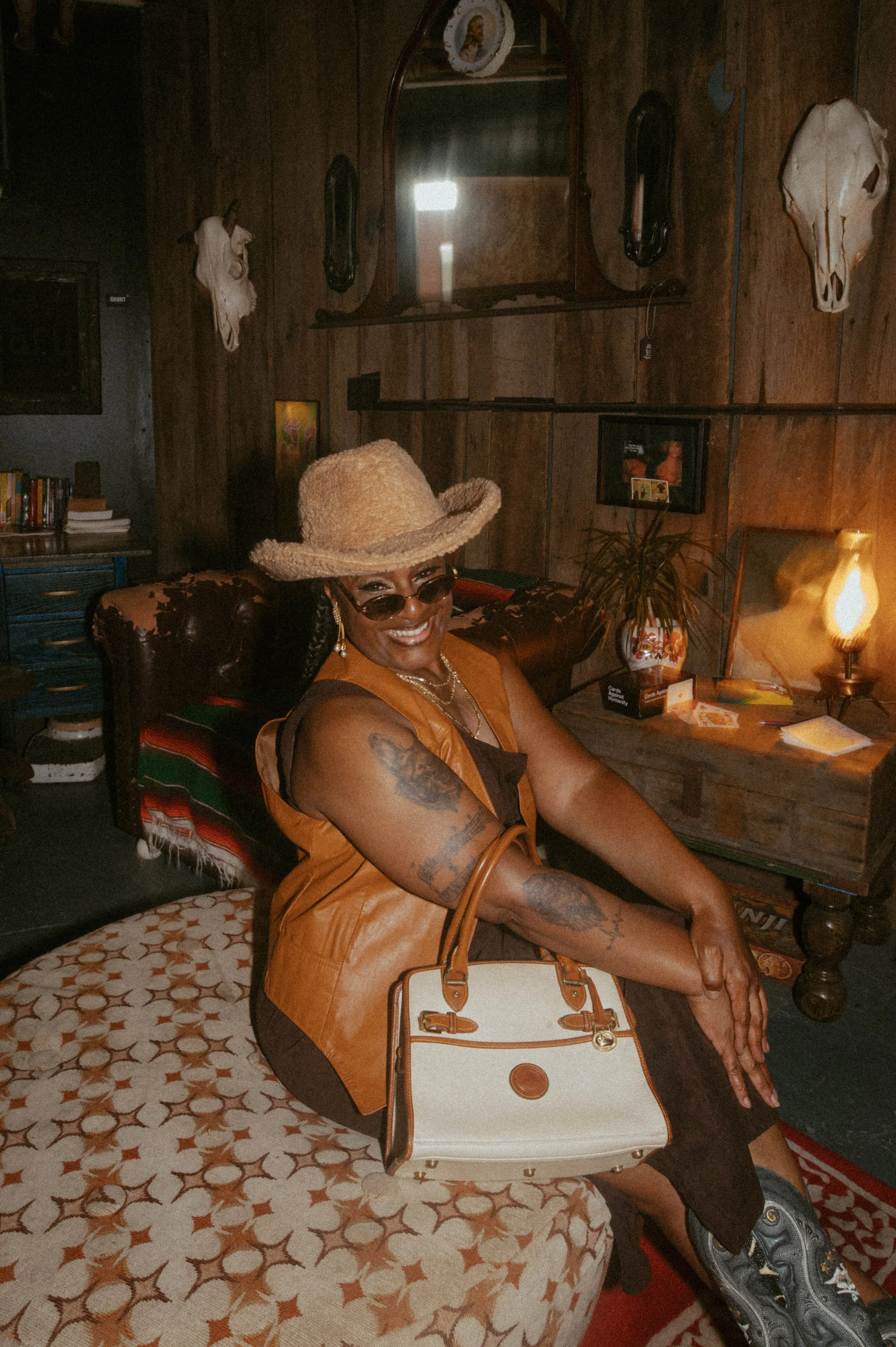 Smiling woman with tattoos, sunglasses, and hat sitting on a patterned chair in a cozy, rustic room with wood-paneled walls, animal skulls, and vintage decor.