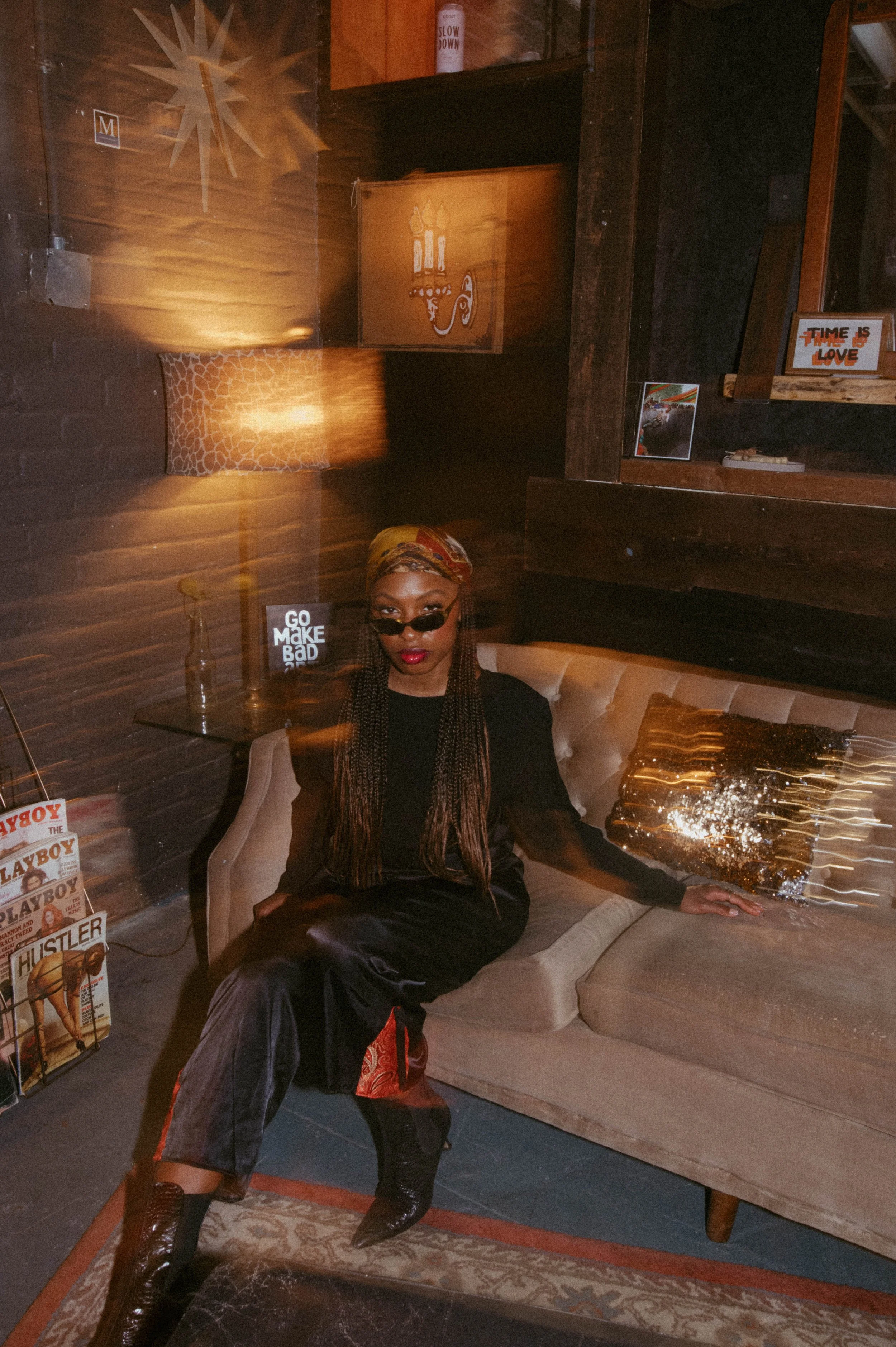 Katana in sleek black outfit with headscarf and sunglasses seated on a vintage sofa in a moody lounge setting, styled with retro magazines and warm ambient lighting, capturing an editorial NYC nightlife aesthetic.