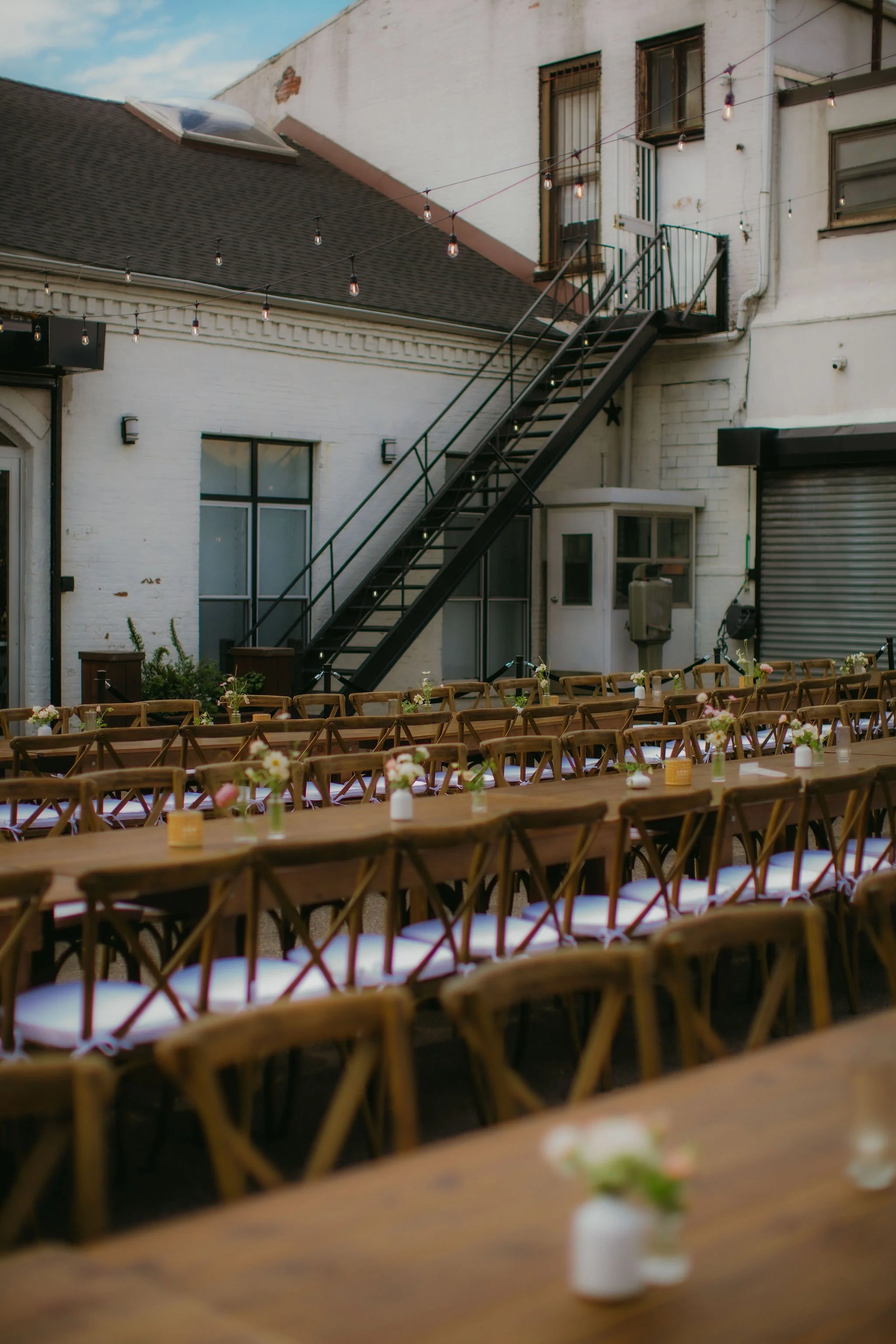 Outdoor event space with long wooden tables and chairs, decorated with small vases of flowers, set against the backdrop of a brick building with fire escape stairs and string lights.