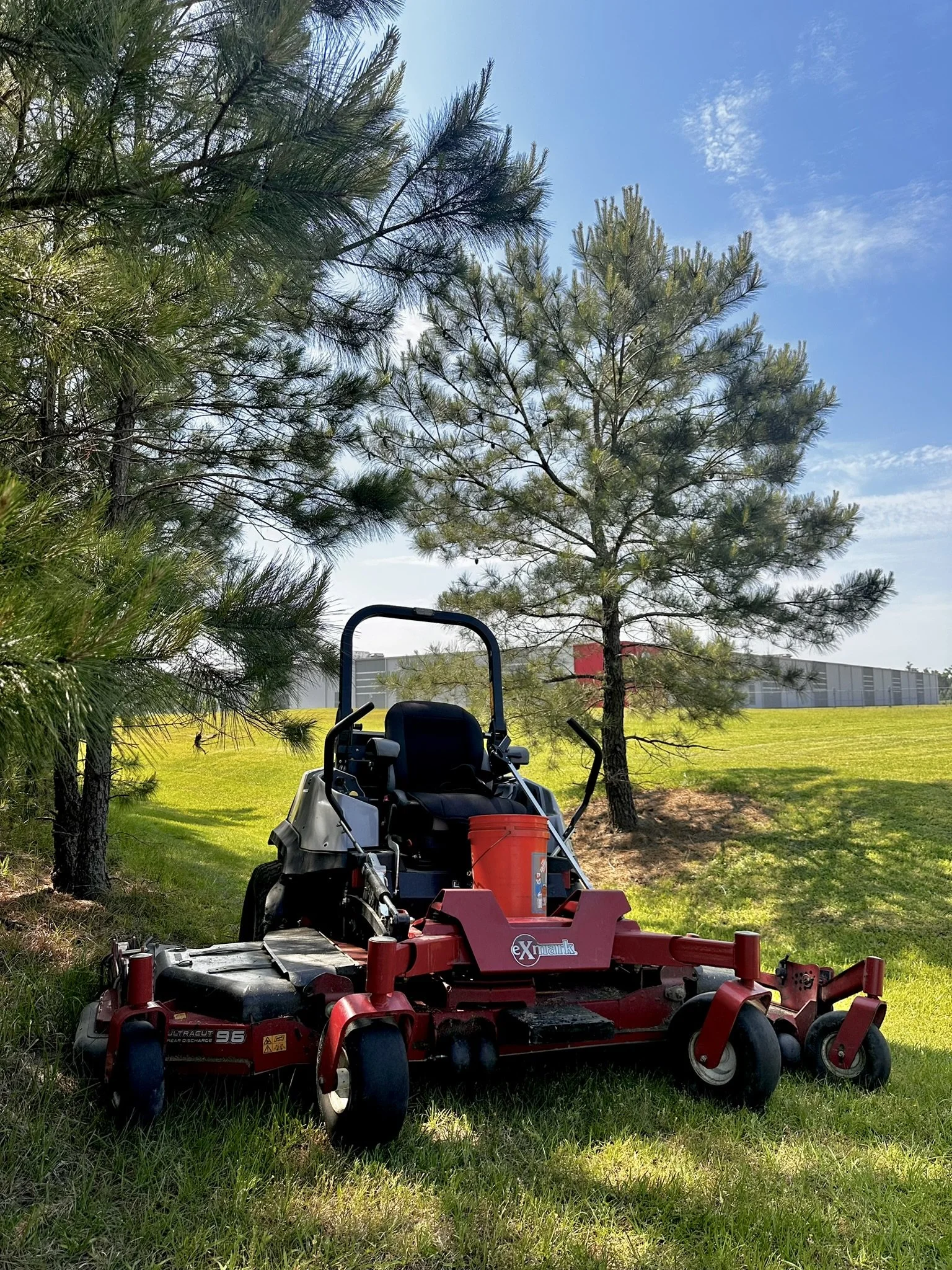 Red commercial mower parked on grass under trees with a building in the background.