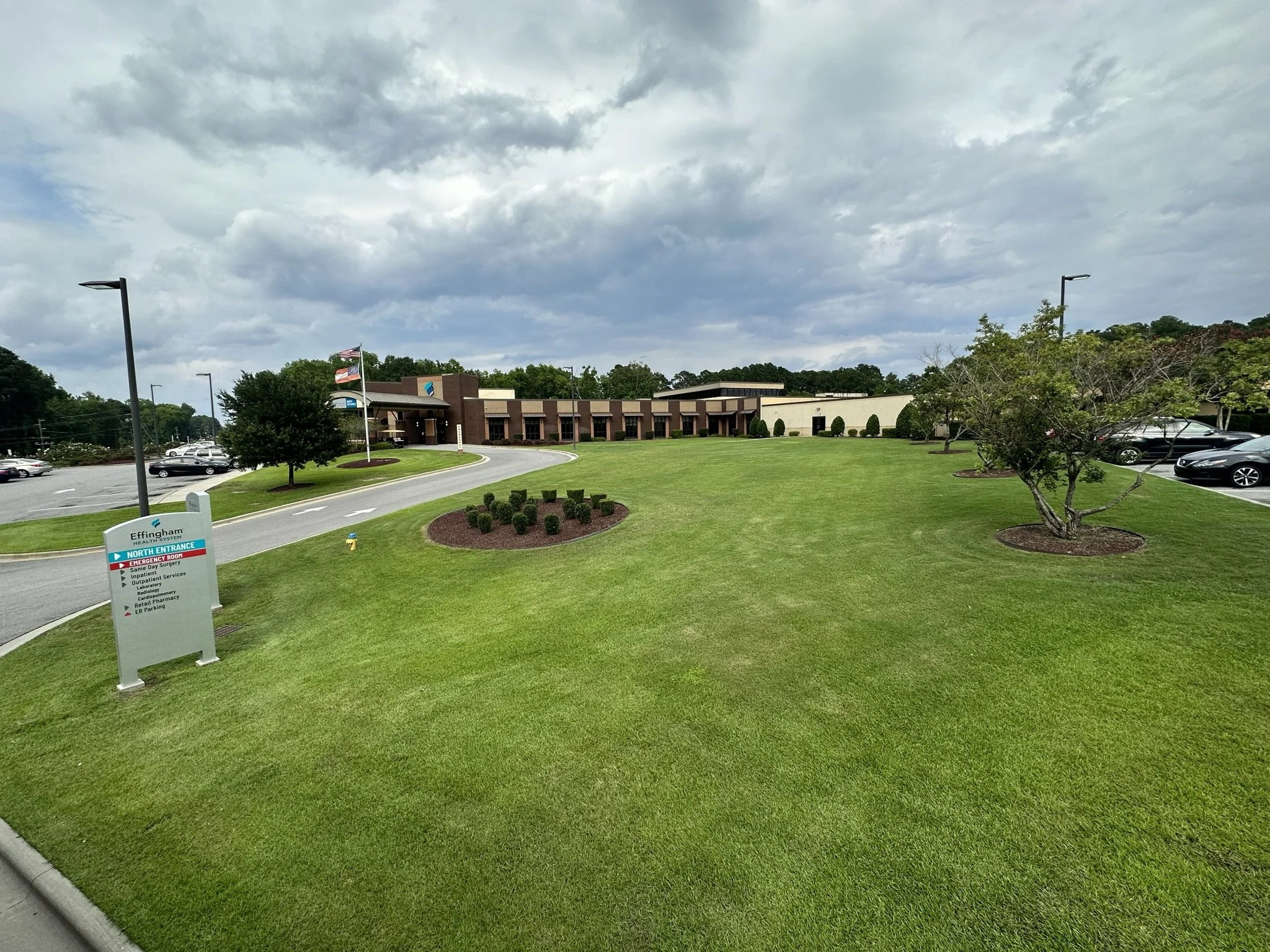 A hospital or healthcare facility with a green lawn, multiple parking lots, trees, and a sign near the entrance. The sky is cloudy with patches of blue.