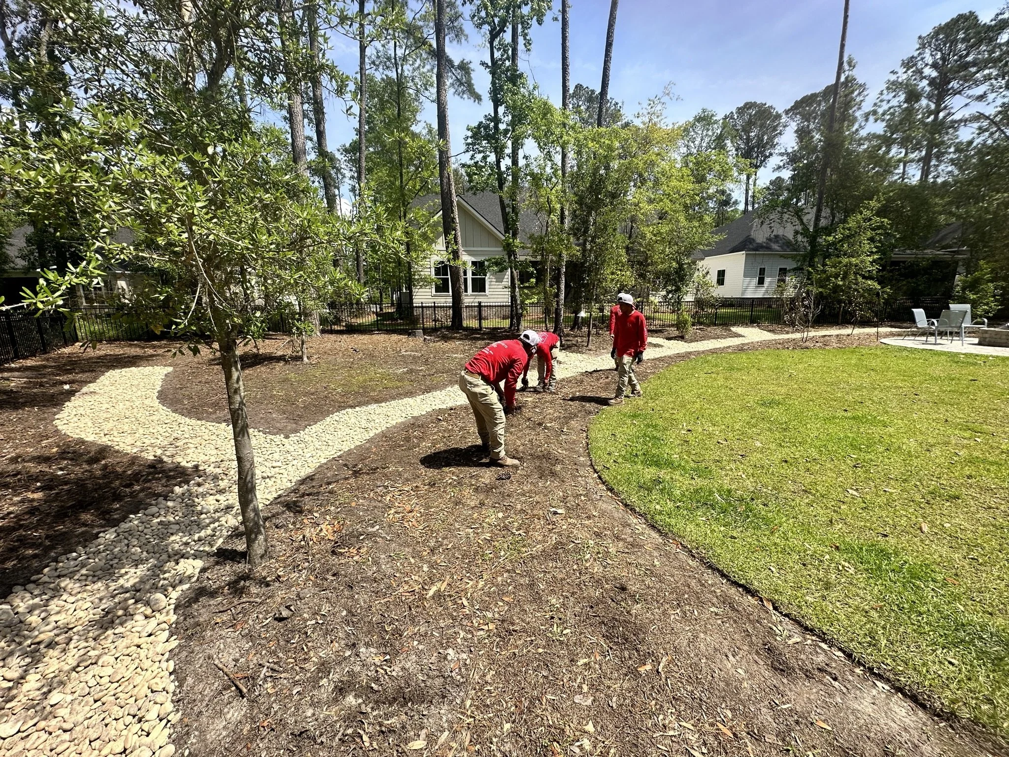 Three workers in red shirts and khaki pants planting a garden in a backyard with a curved stone pathway, grass, trees, and houses in the background.
