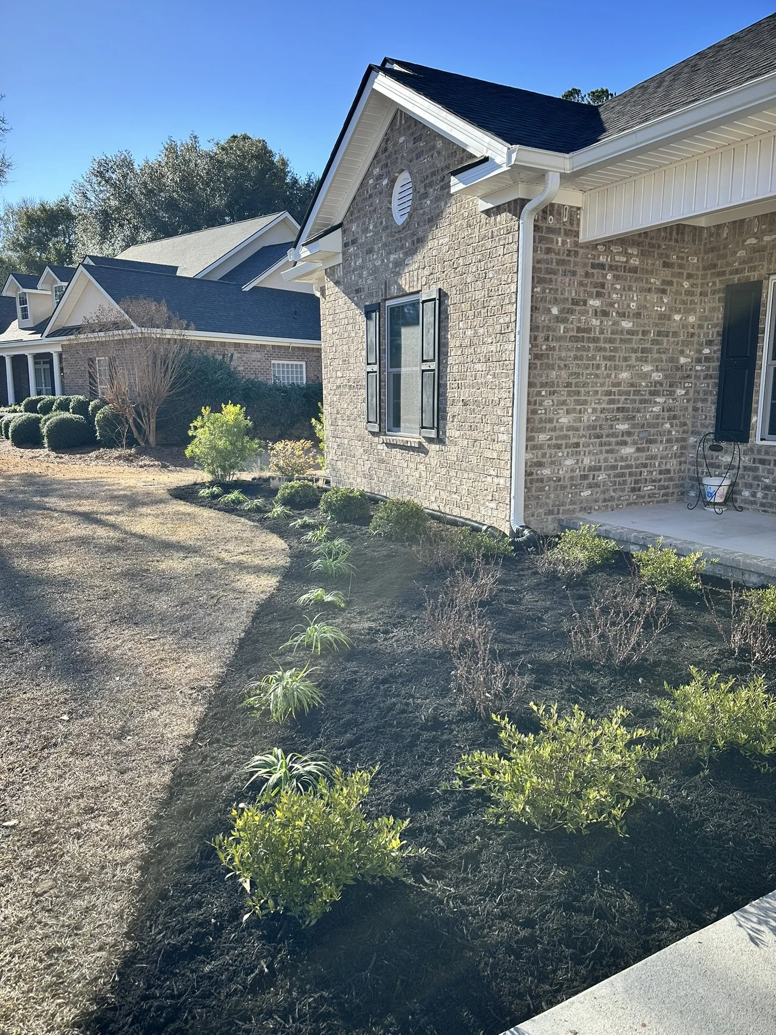 Front yard with newly planted shrubs and flowers, house with brick exterior, black shutters, and a porch, and neighboring houses in background under blue sky.