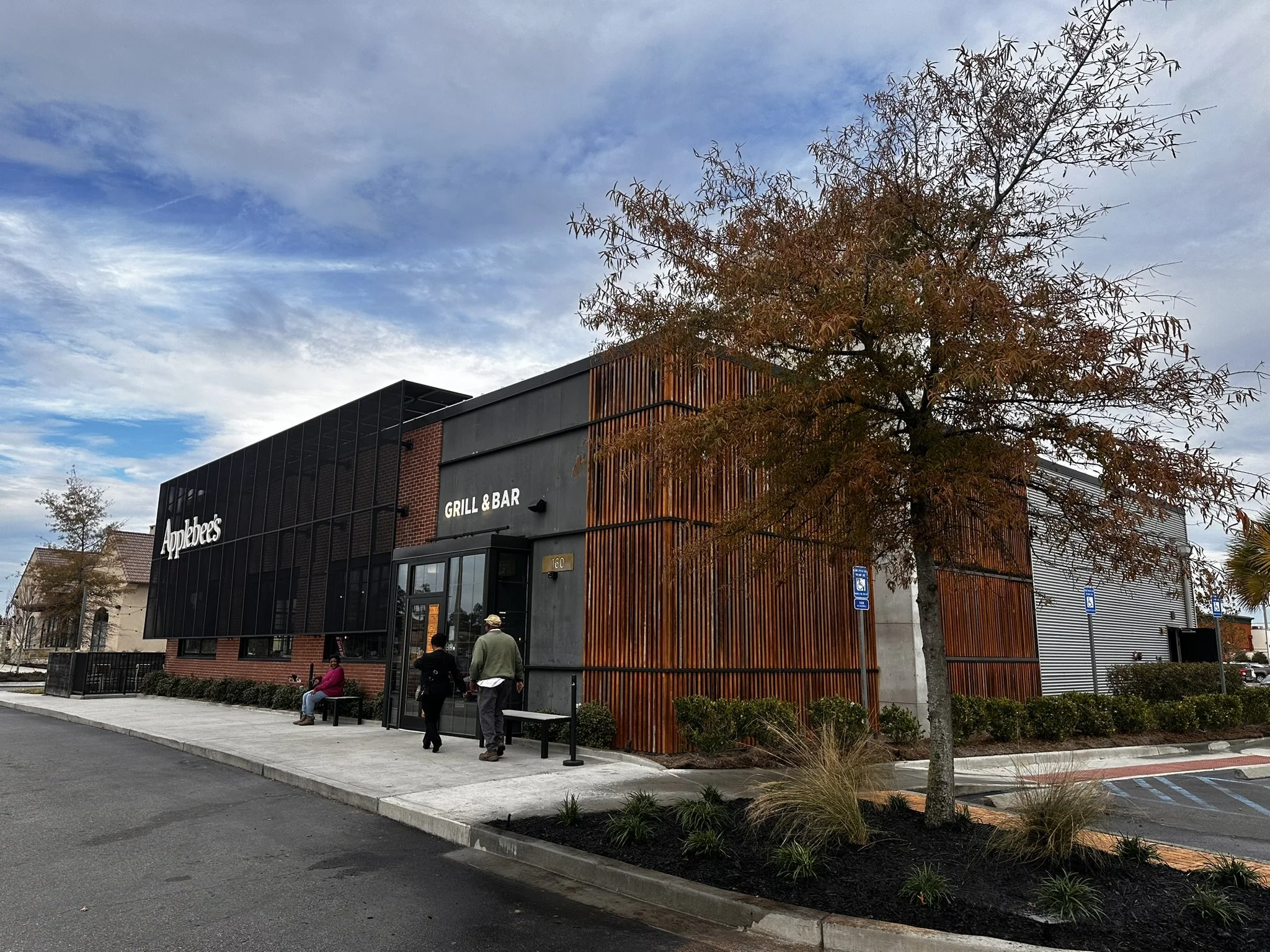 Exterior of an Applebee's restaurant with a modern design, featuring wood and metal siding, with a few people sitting and walking outside, and parking spaces visible nearby.