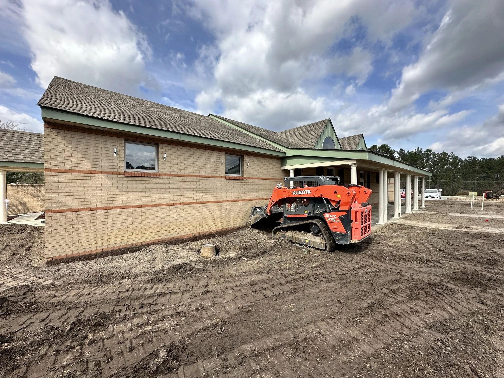 Construction site showing a partly built brick building with a green roof and pillars, and a small orange Kubota skid steer loader on dirt ground under a cloudy sky.