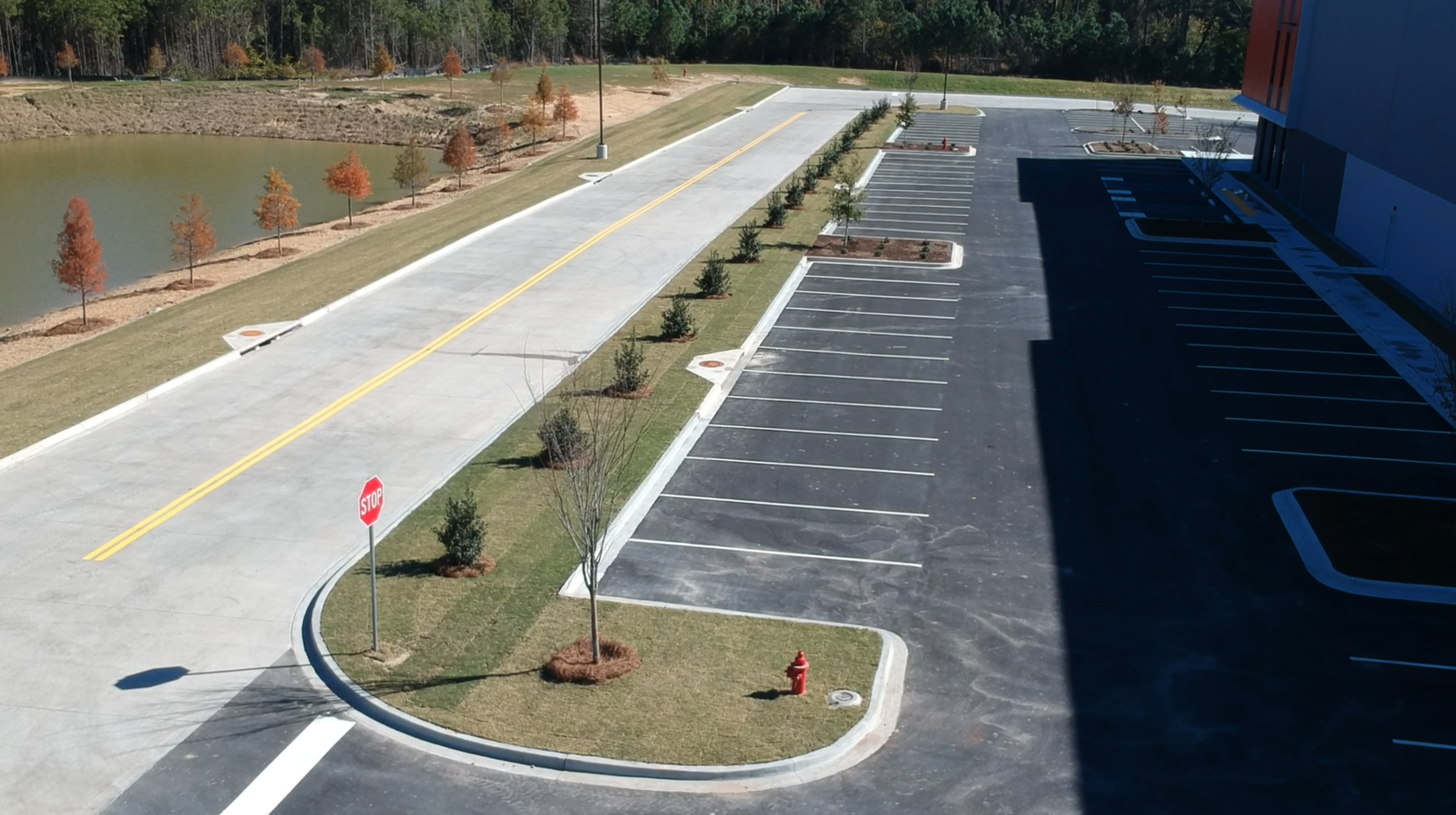 Empty parking lot next to a small body of water with trees and some landscaping, a fire hydrant, and a stop sign at the entrance.