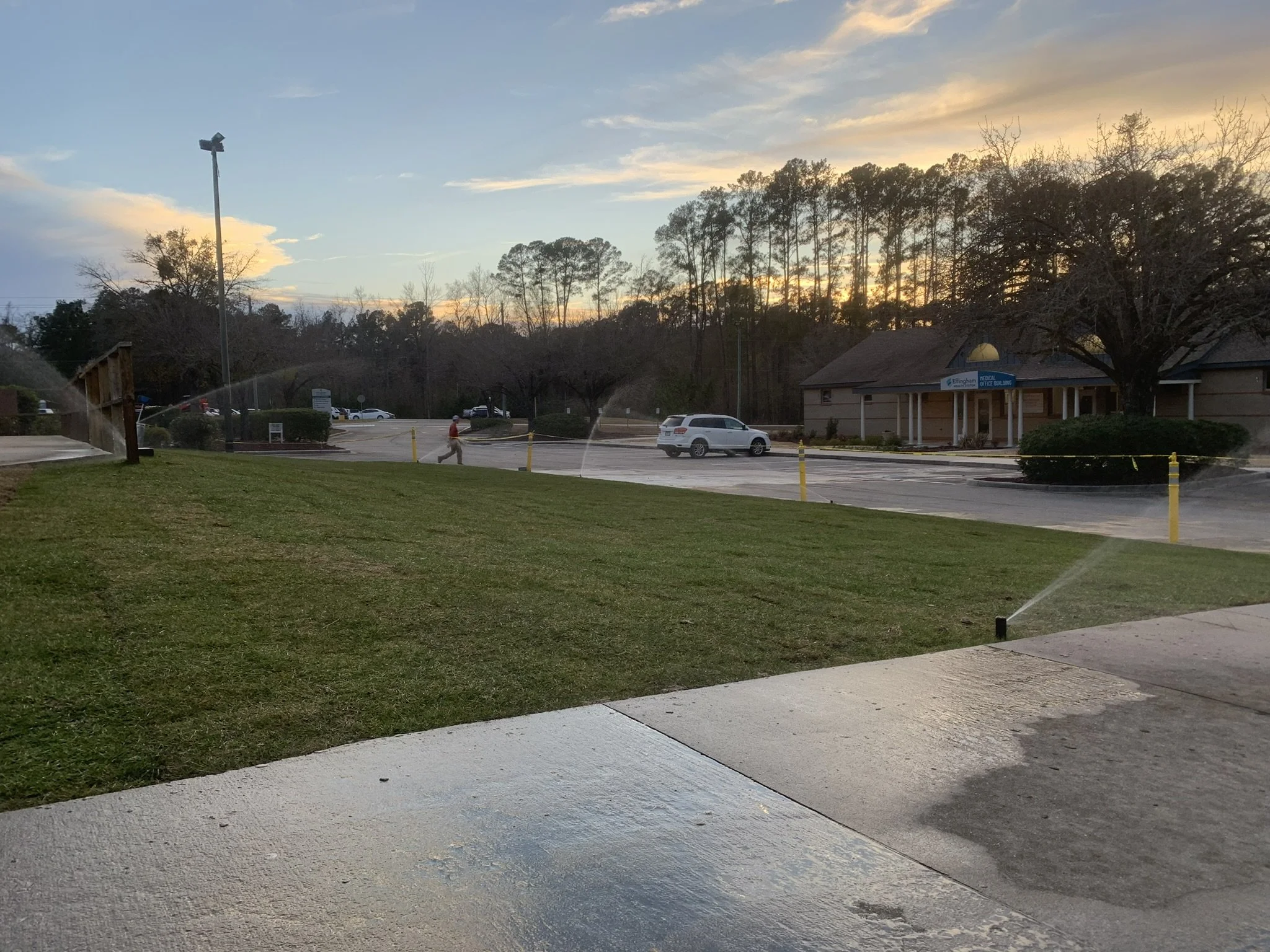 Sunset over a parking lot with sprinklers watering the grass in the foreground, a building on the right, and trees in the background.