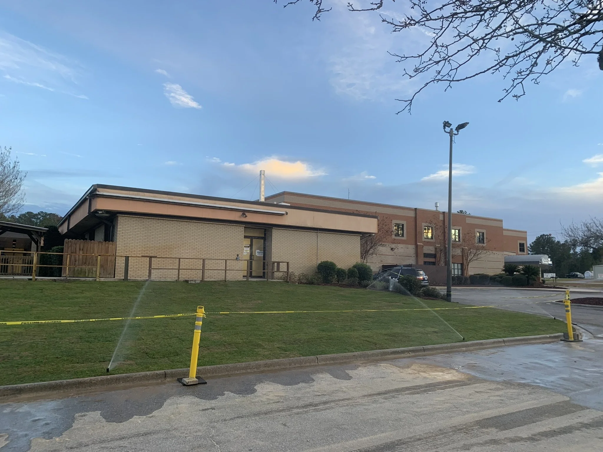 A low-rise building with a brick and beige exterior, surrounded by a grassy lawn with sprinklers watering the grass. There are some shrubs and trees nearby, with a parking lot and cars in the background. The sky is partly cloudy with blue skies.