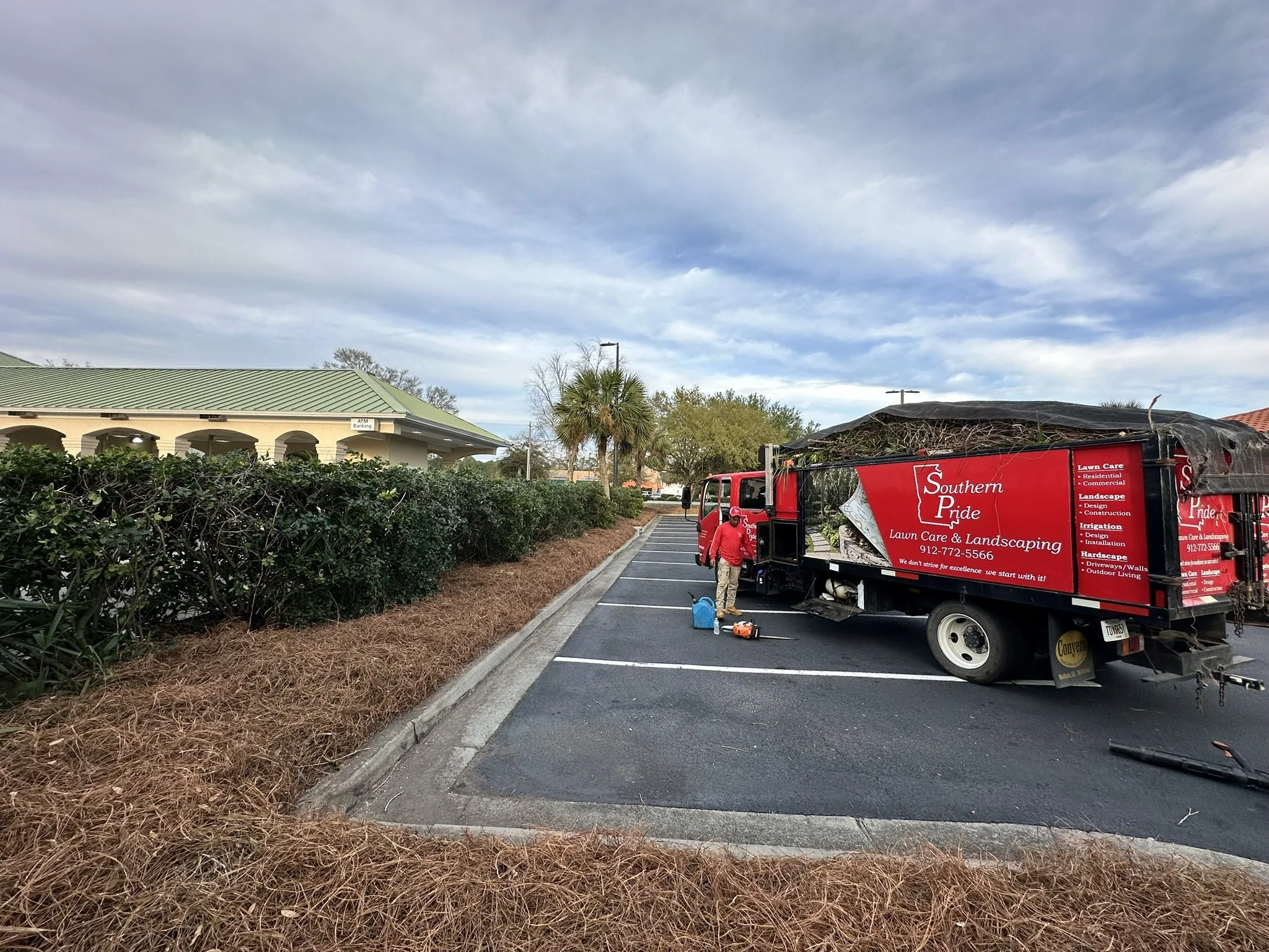 Lawn care and landscaping truck parked in a parking lot with workers preparing equipment, with bushes and a building with green roofing in the background, under a cloudy sky.