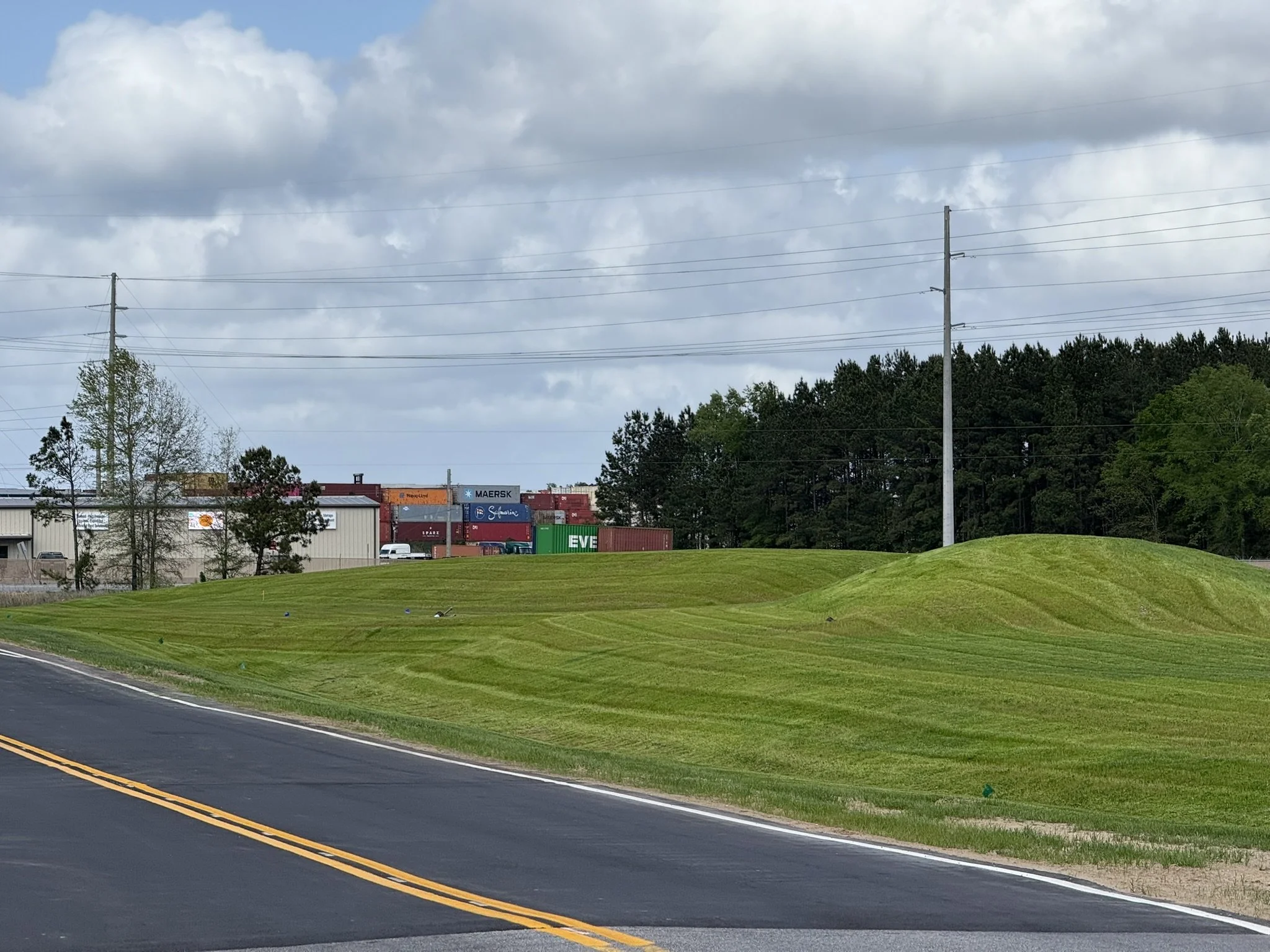 Well-manicured grassy area next to a paved road with yellow lines, with trees, utility poles, and a cluster of shipping containers in the background under a cloudy sky.