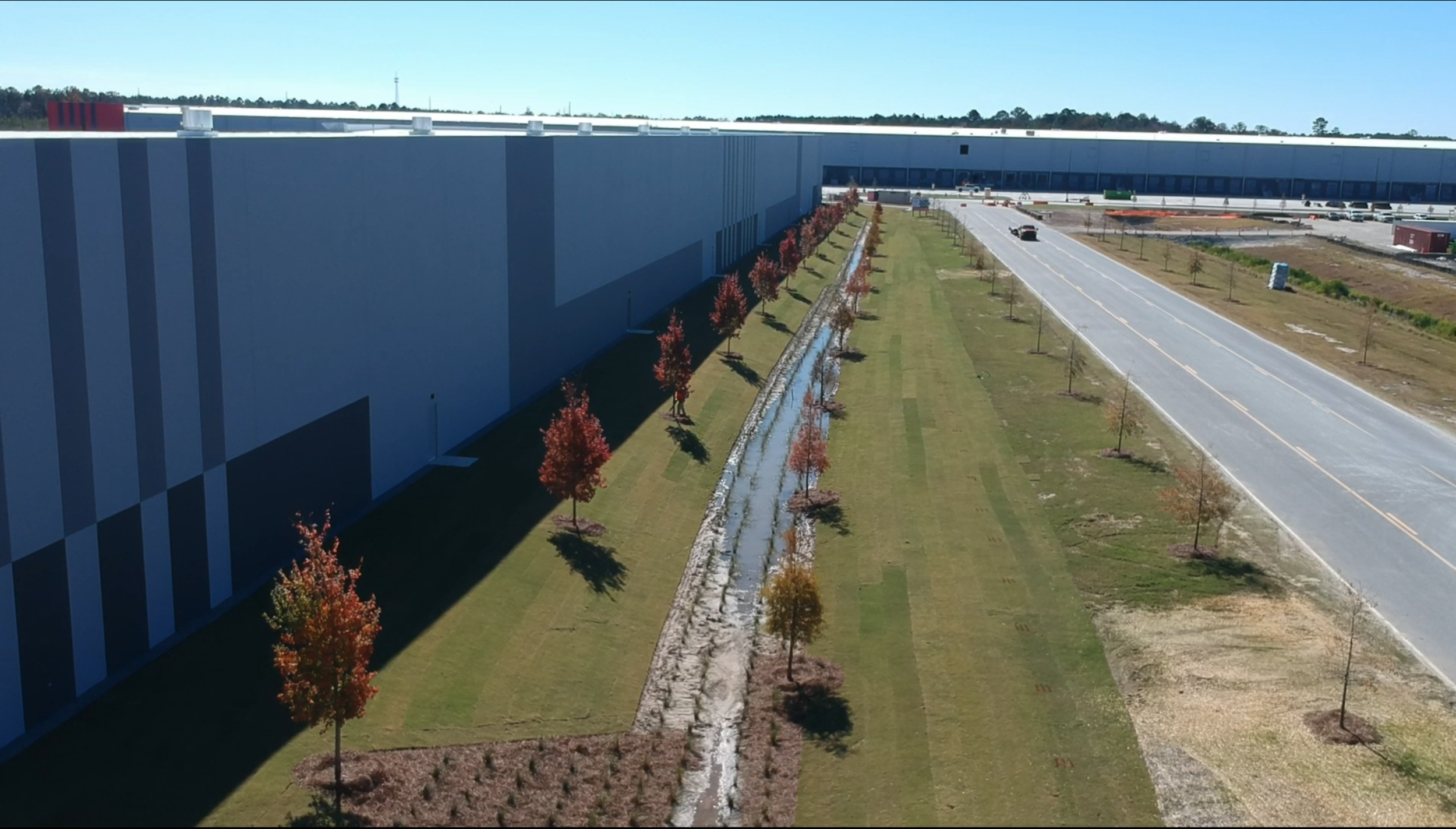 A large industrial or commercial building with a landscaped yard and a narrow water feature running alongside the building, trees with fall foliage, and a road in the background under a clear blue sky.