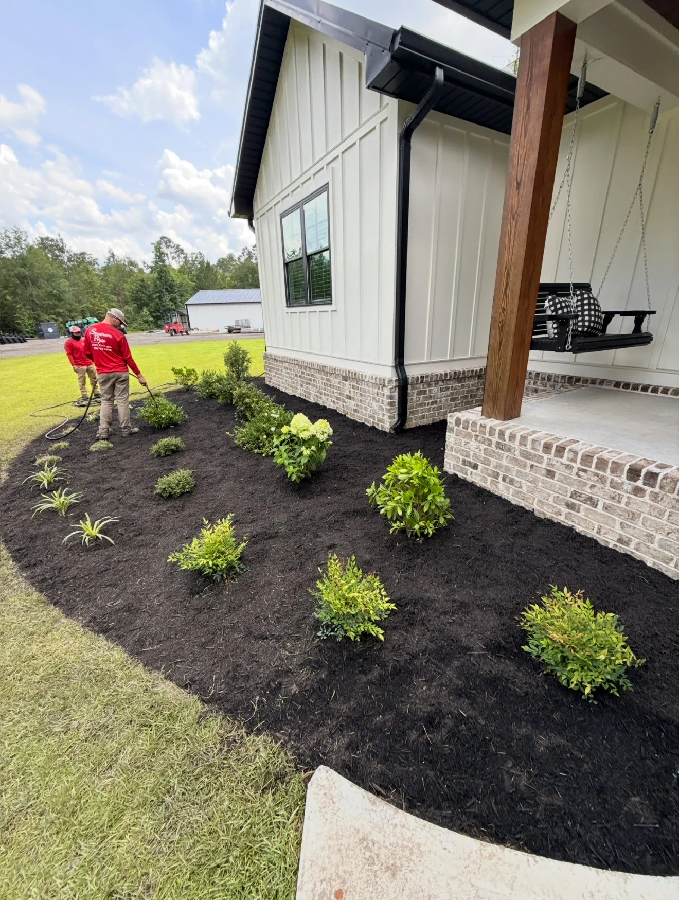 Two people wearing red shirts planting bushes in a freshly mulched garden bed next to a house with white siding and brick foundation, under a partly cloudy sky.