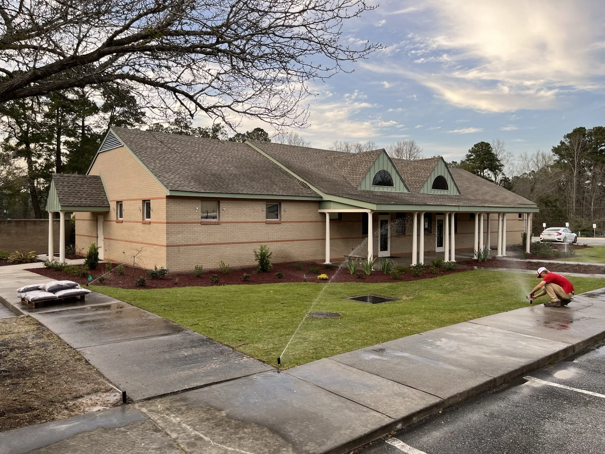 A worker watering the front lawn of a beige brick building with a grey roof and green accents, situated by a parking lot with some cars and trees in the background on a partly cloudy day.