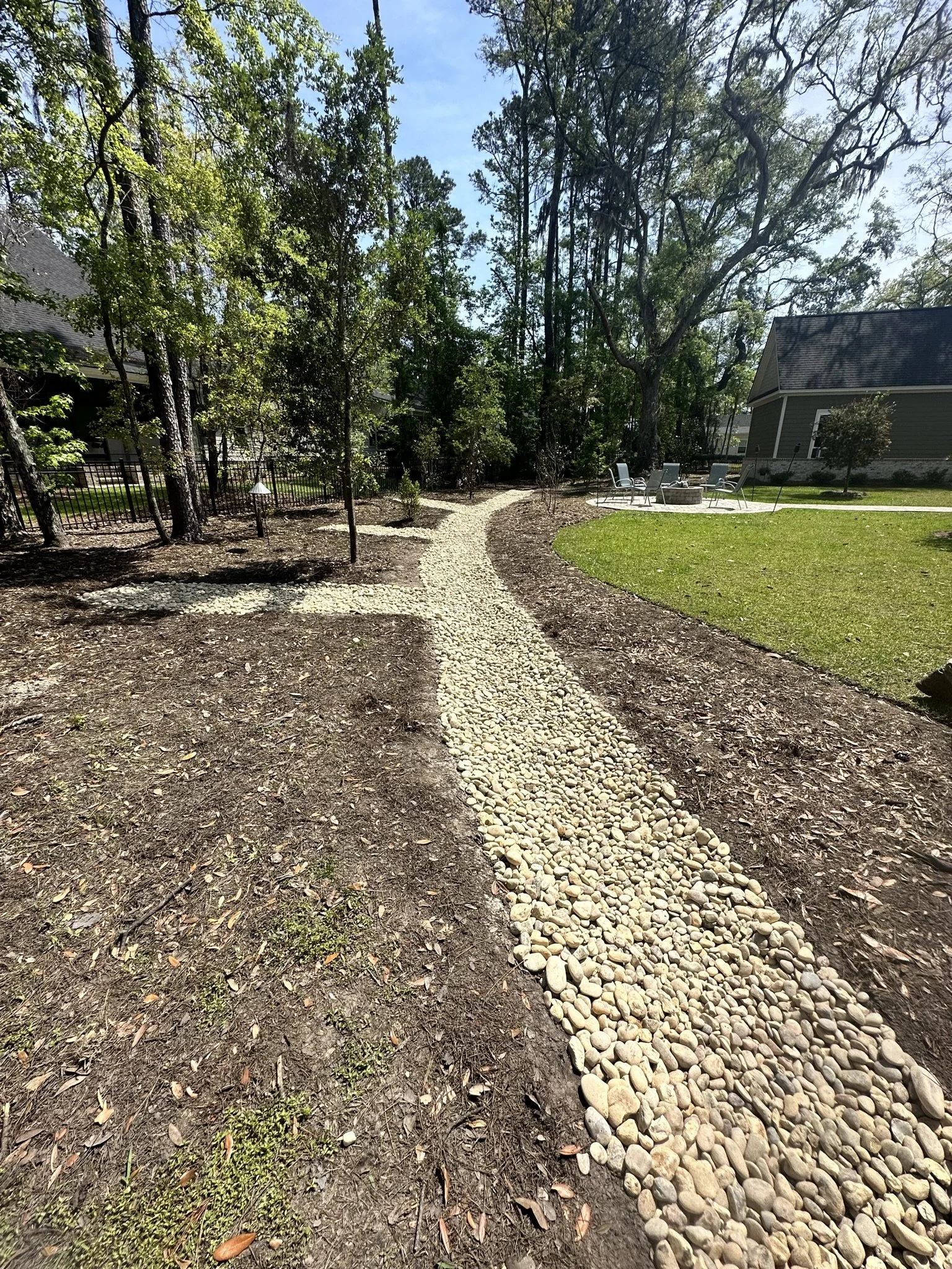 Curved pebble pathway through a yard with grass, trees, and outdoor seating in the background.