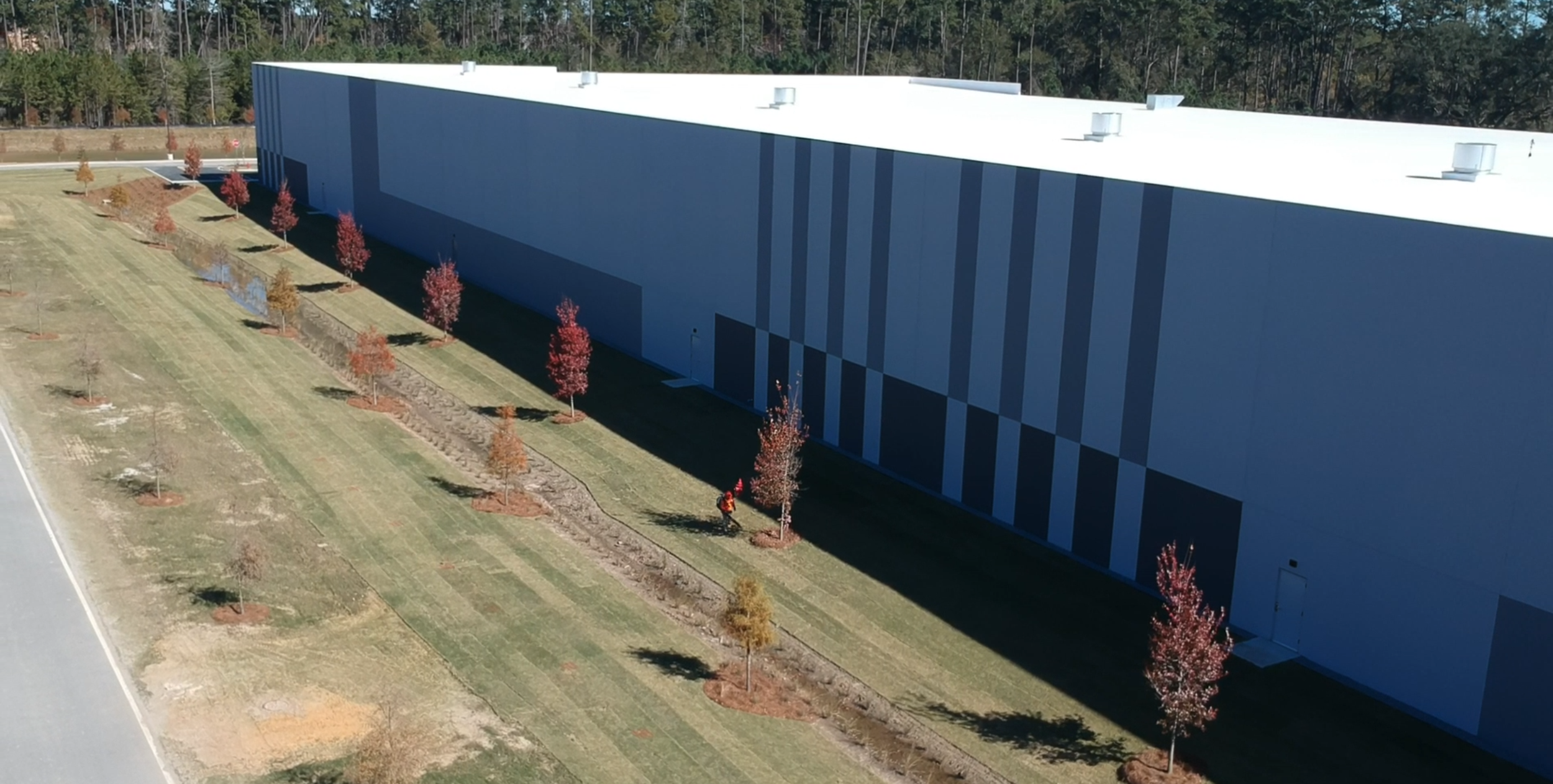 A large industrial warehouse building with a white roof, surrounded by a landscaped area with small trees and grass, and a road at the edge of the image.